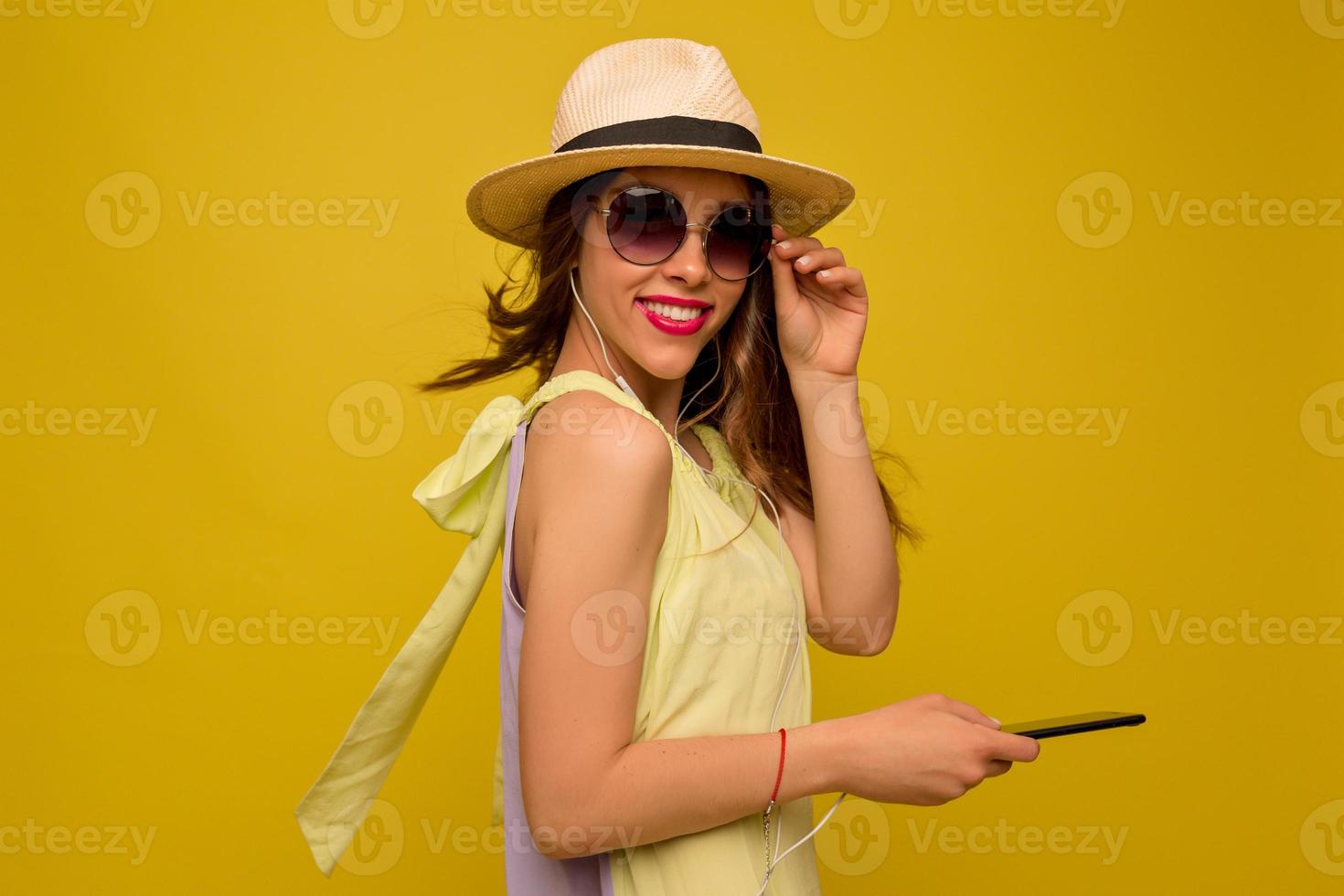 affascinante donna alla moda che indossa occhiali e cappello girarsi alla telecamera con lo smartphone su sfondo giallo isolato e sorridente, vacanza, viaggio, emozioni felici foto