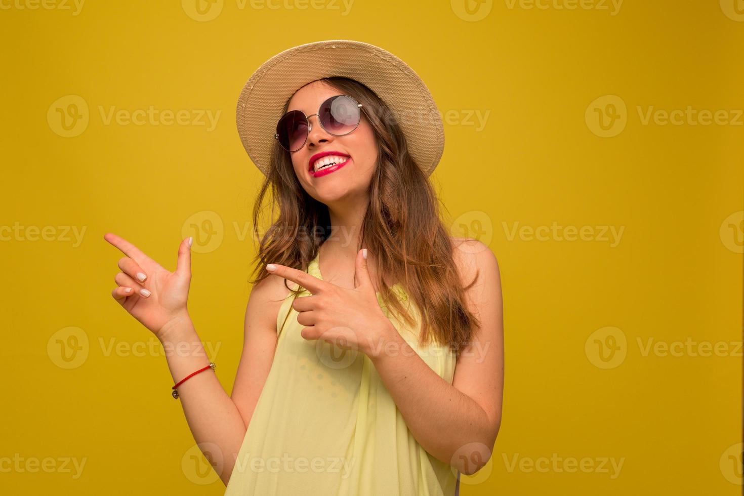 felice donna bruna in abito giallo e cappello guarda in alto, puntando il dito e sorride sinceramente su sfondo giallo. foto