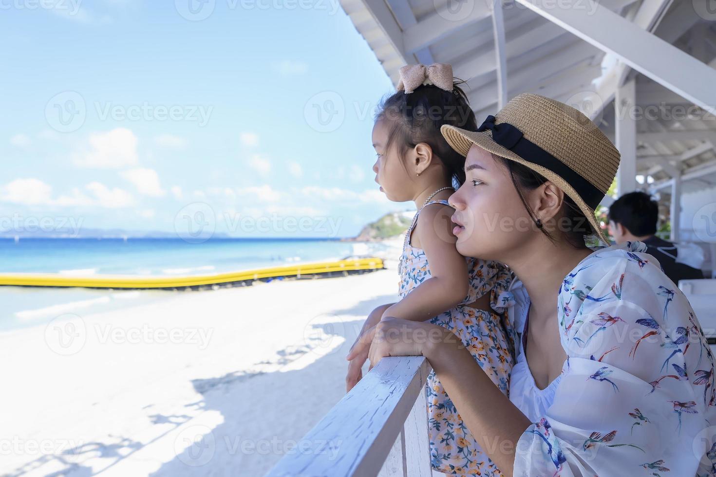 felice figlia e madre seduta al ristorante vicino al mare e guardando la splendida vista della spiaggia tropicale. persone che guardano il cielo e il mare blu. stile di vita di viaggio durante le vacanze estive in famiglia con i bambini foto