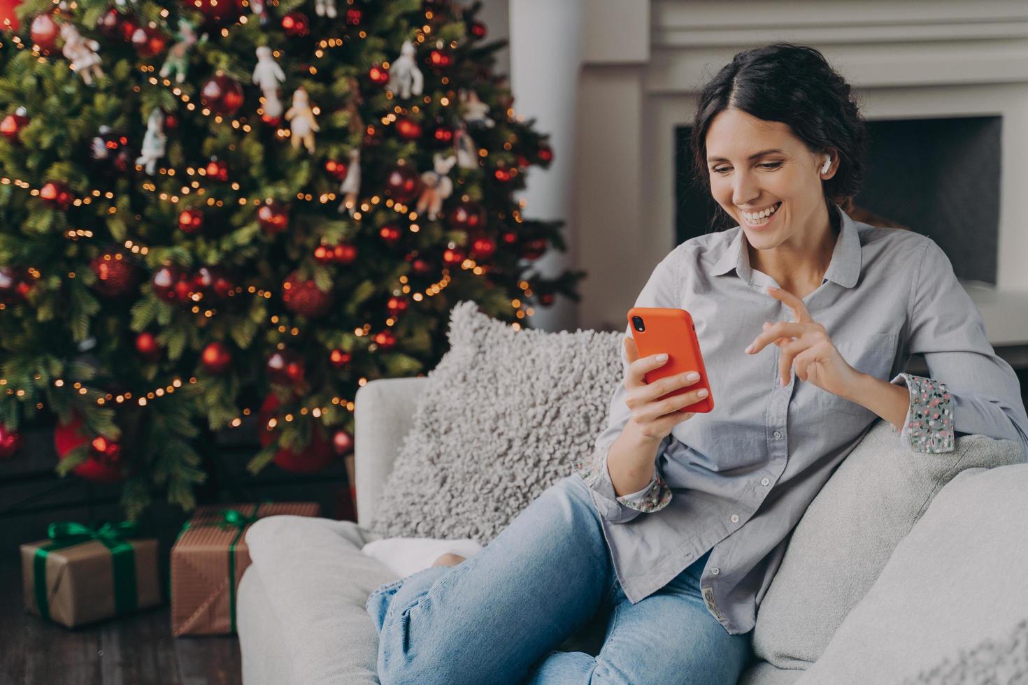 femmina felice in auricolari che tiene smartphone, parla con la famiglia o gli amici online durante il periodo natalizio foto