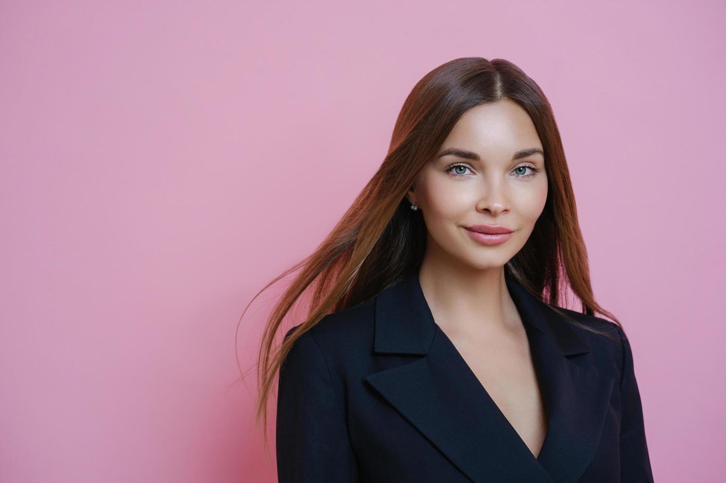 ritratto di una splendida manager femminile che va al lavoro, indossa un costume formale nero, trucco naturale, sembra sicuro di sé alla telecamera, si erge su sfondo rosa dello studio. persone e concetto di cura. foto