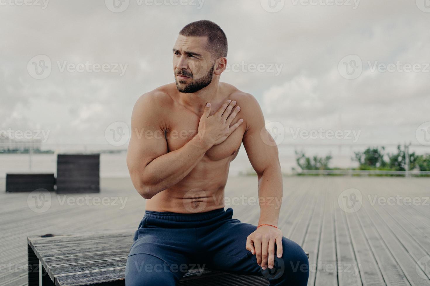 l'uomo barbuto premuroso si riposa dopo un intenso allenamento cardio, tiene la mano sul petto e ha mancanza di respiro, si siede all'aperto, concentrato da parte, posa in topless. sport e stile di vita sano foto