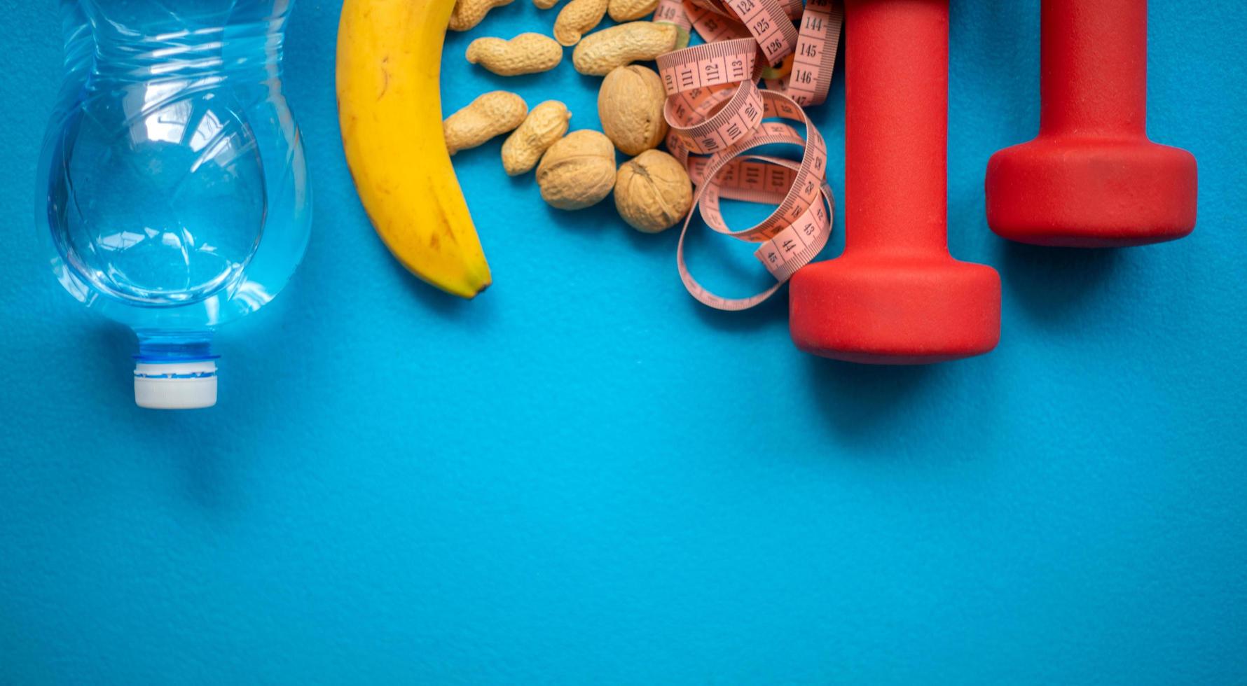 banana, noci, acqua, metro a nastro e manubri su sfondo blu. cibo e attrezzature per il fitness per uno stile di vita sano foto
