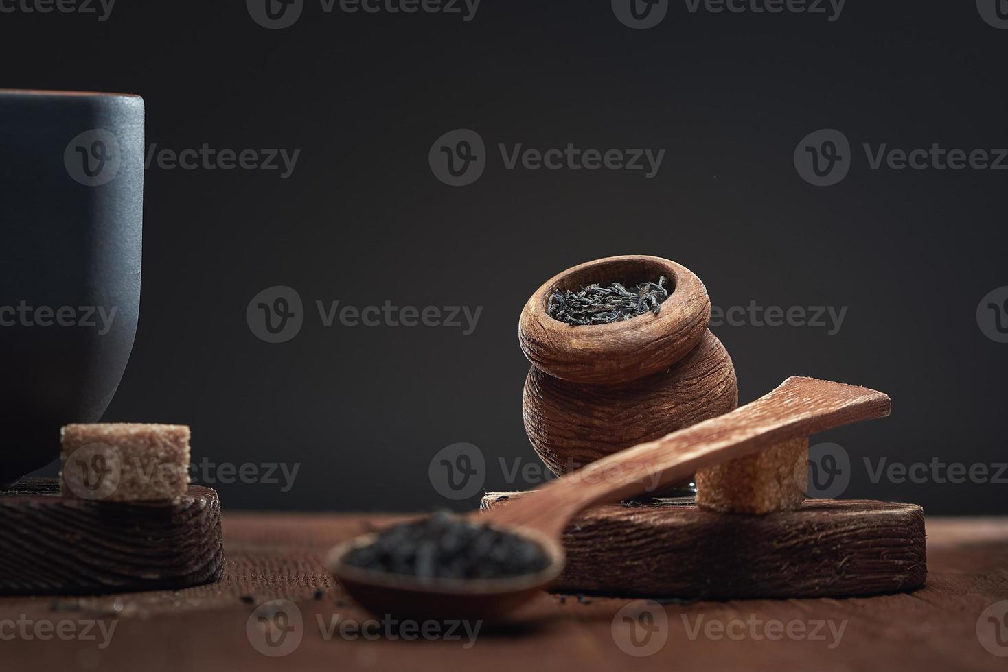 tè secco in cucchiai di legno e in un vasetto di legno e pezzetti di zucchero di canna sul tavolo, su sfondo scuro. foto