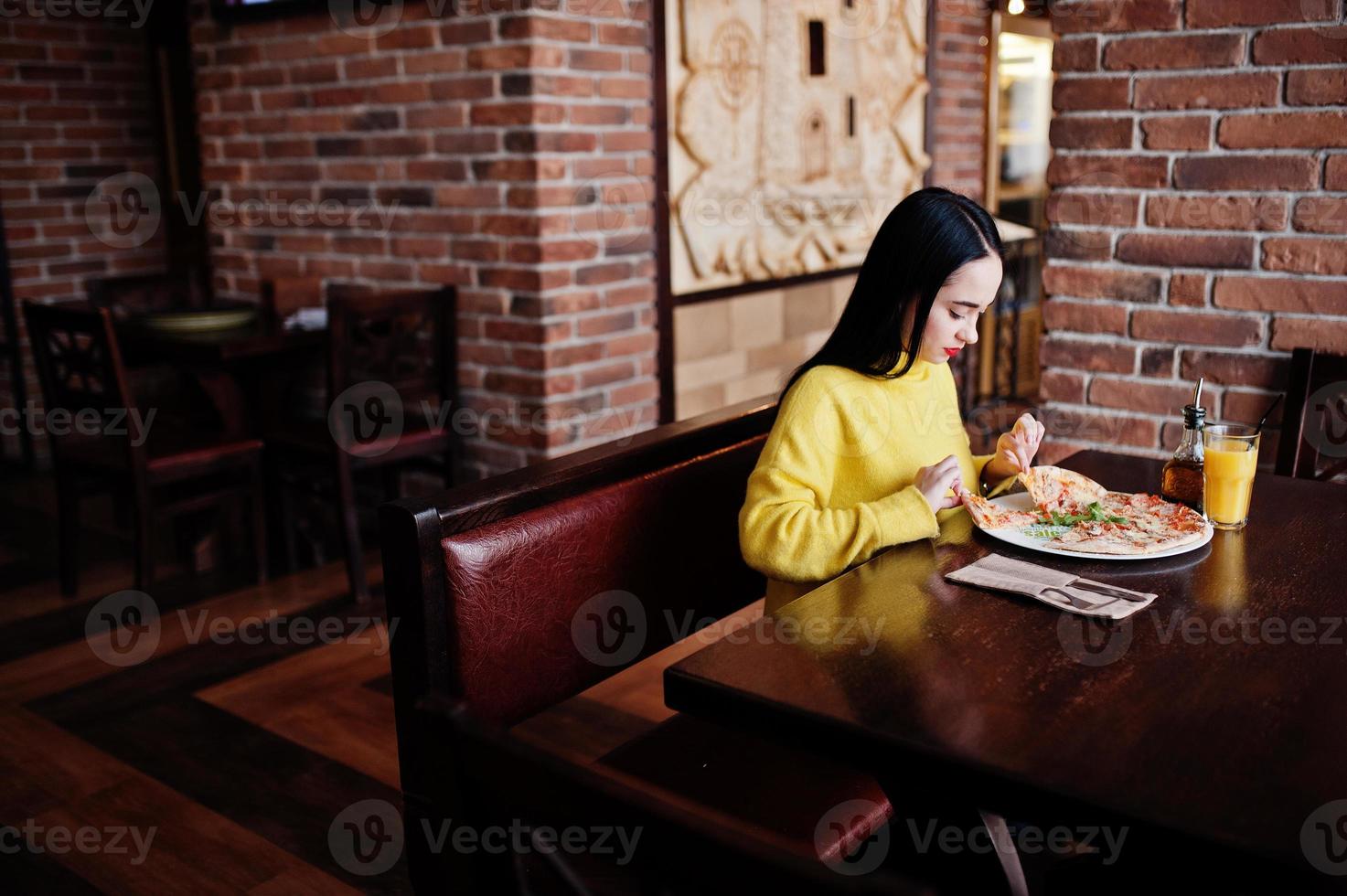 divertente ragazza bruna in maglione giallo che mangia pizza al ristorante. foto