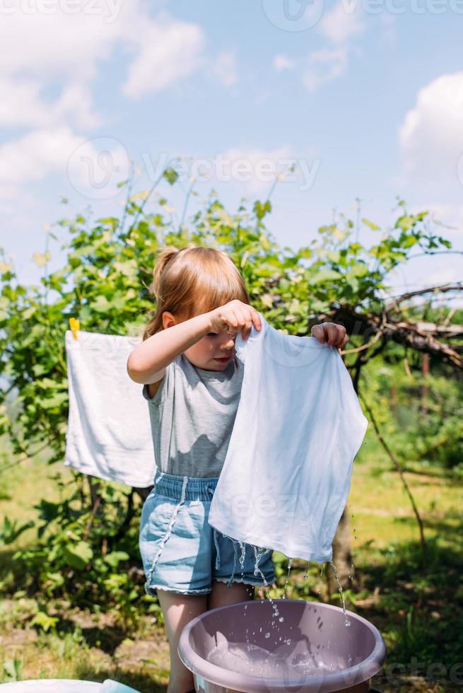 la bambina in età prescolare aiuta con il bucato. il bambino lava i vestiti in giardino foto