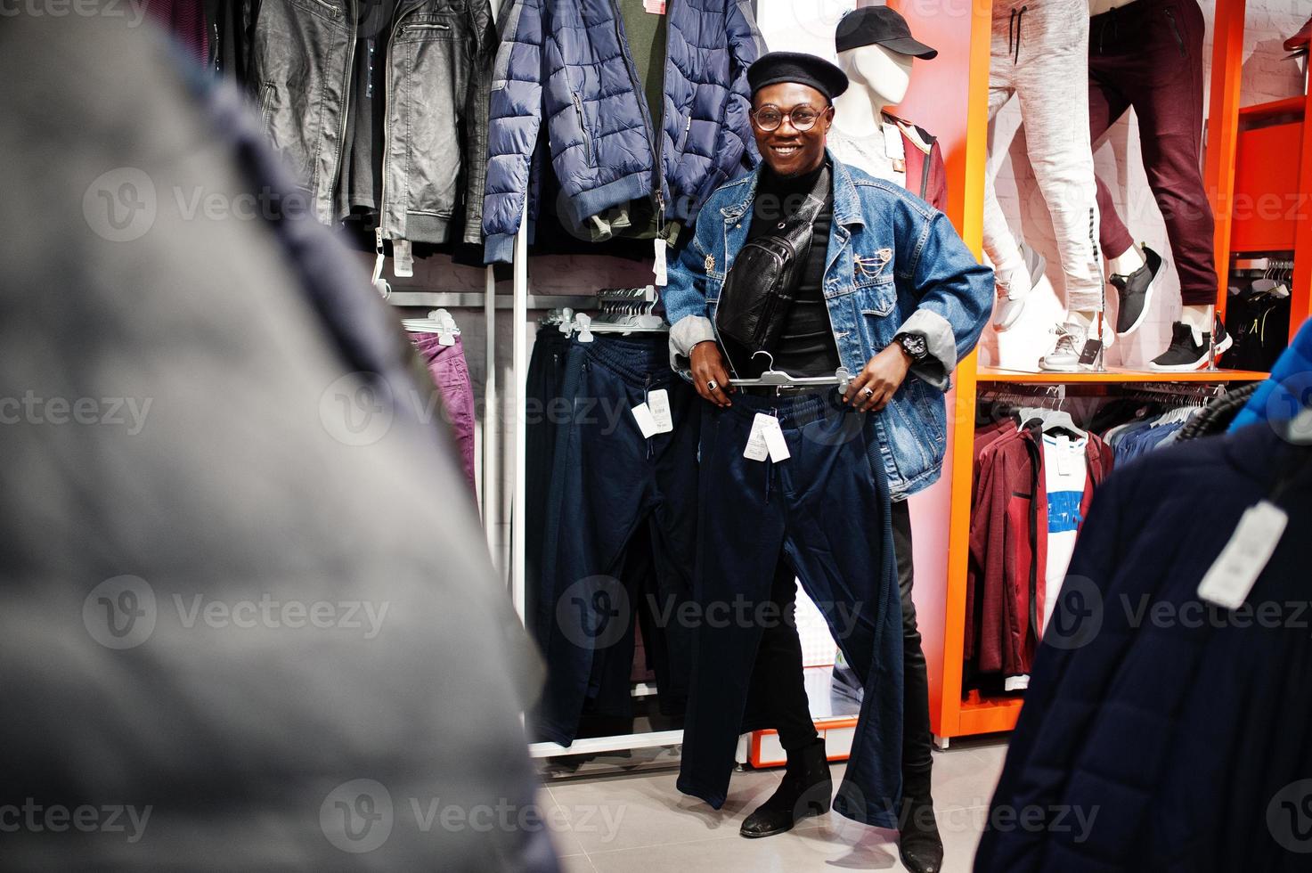 uomo afroamericano casual elegante alla giacca di jeans e berretto nero al negozio di vestiti alla ricerca di nuovi pantaloni sportivi. foto