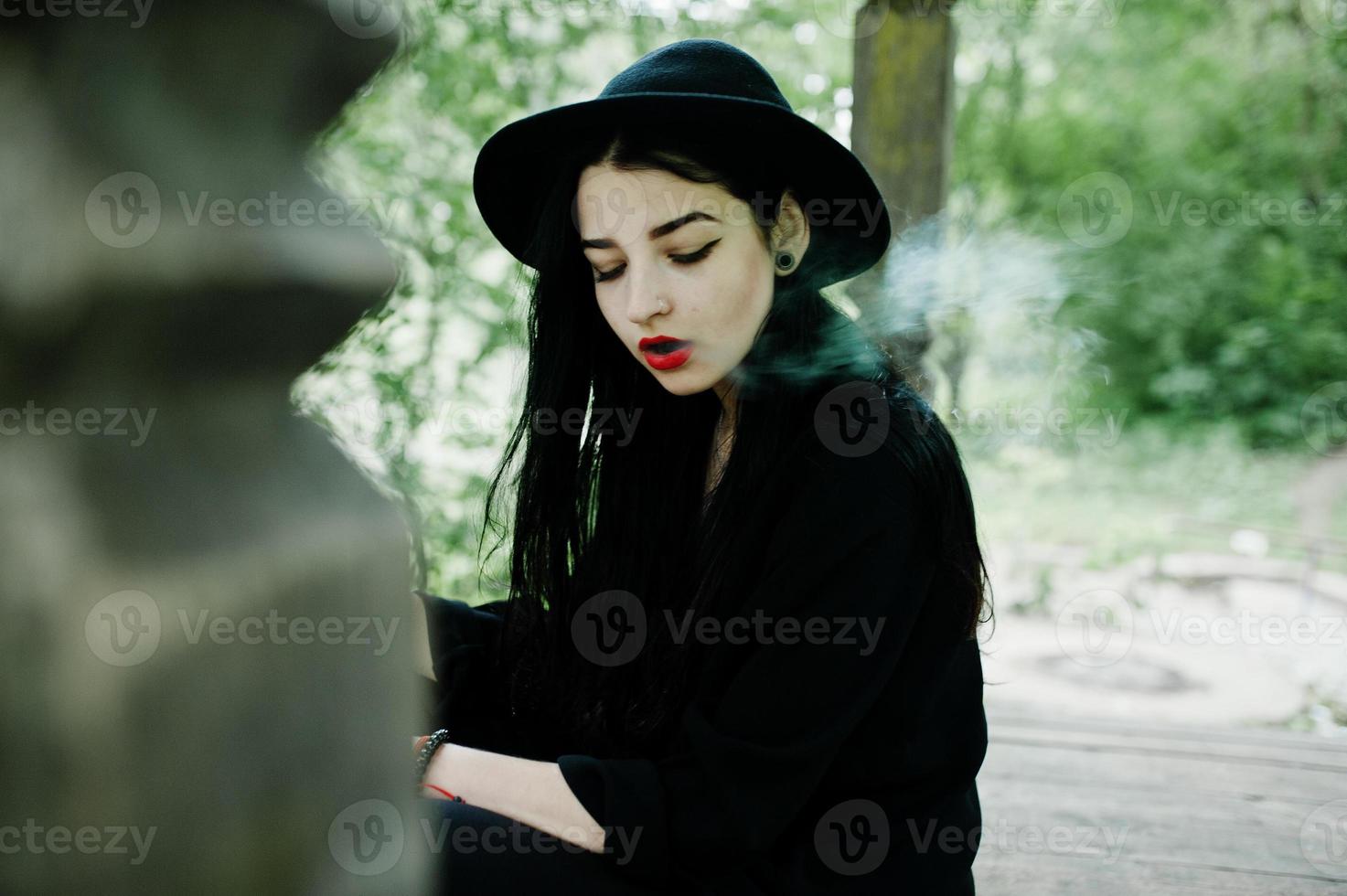 sensuale fumatrice tutta in nero, labbra rosse e cappello. donna drammatica goth che fuma sigaretta sottile. foto