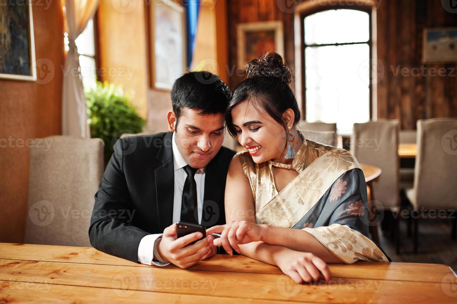 coppia di amici indiani eleganti e alla moda di donna in sari e uomo in vestito posato caffè al coperto e guardando qualcosa sul telefono cellulare. foto