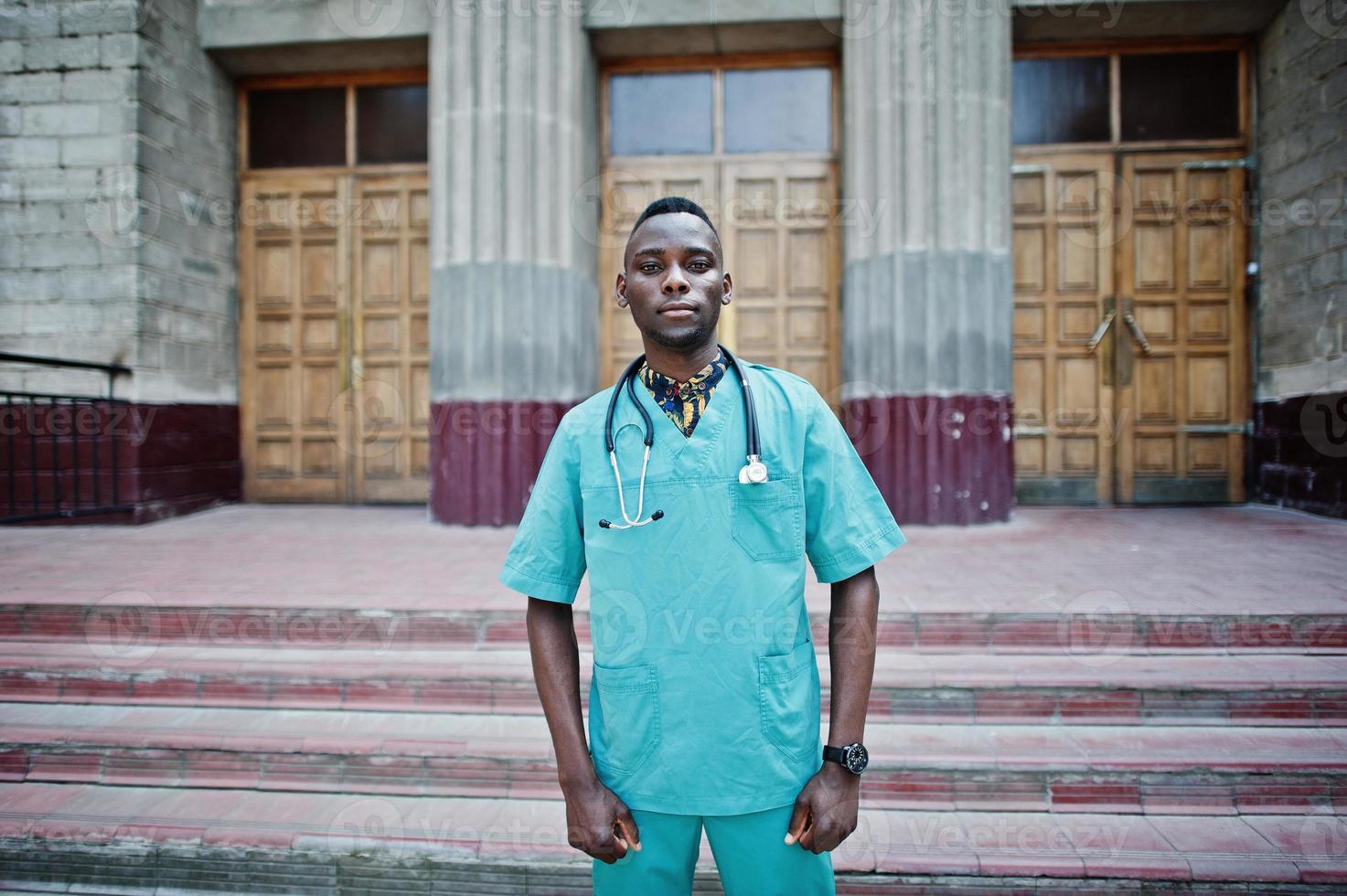 medico afroamericano maschio al camice da laboratorio con lo stetoscopio all'aperto contro la porta della clinica. foto