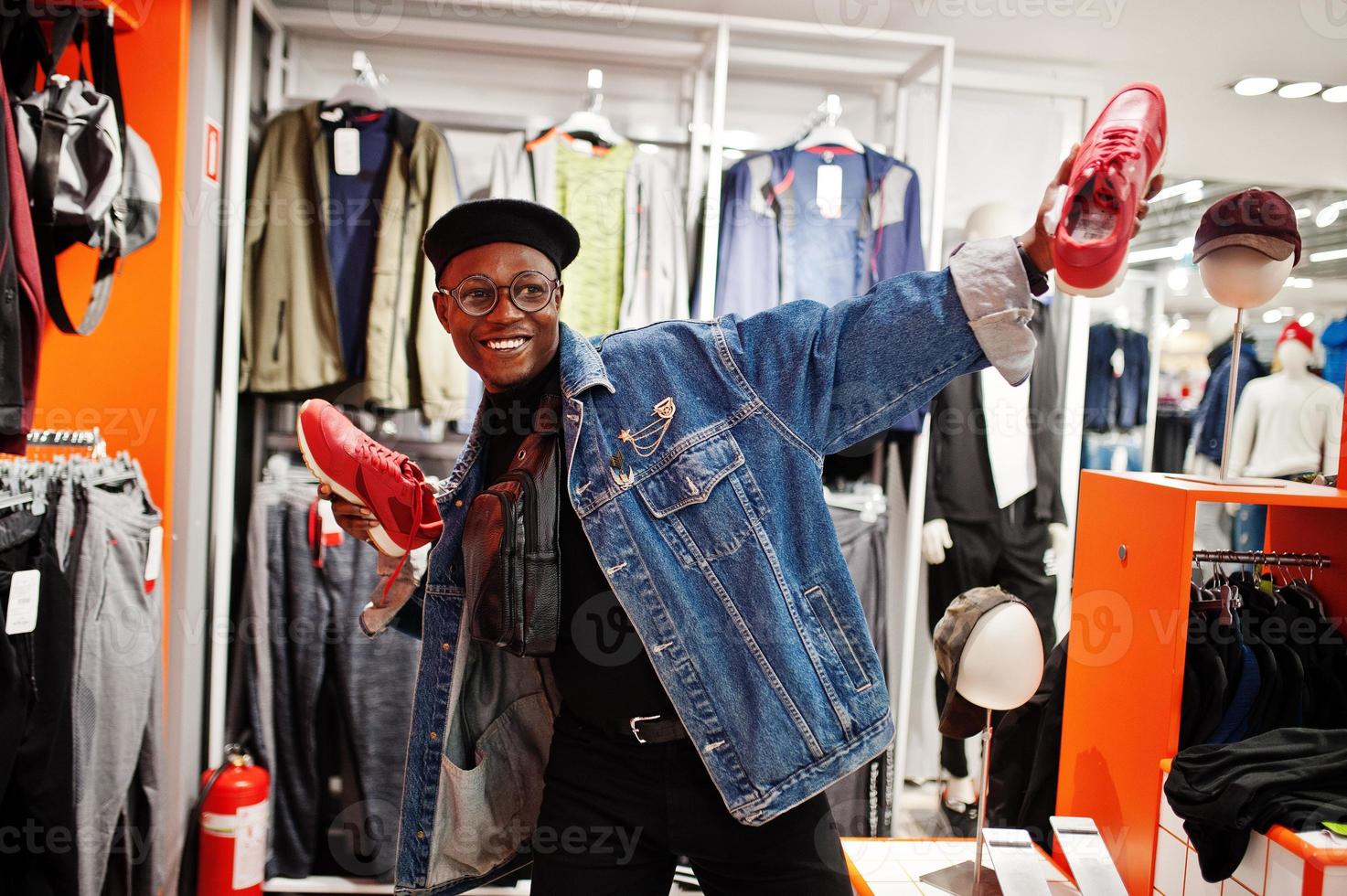 uomo afroamericano casual elegante alla giacca di jeans e berretto nero al negozio di vestiti che tiene nuove scarpe da ginnastica rosse a portata di mano. foto