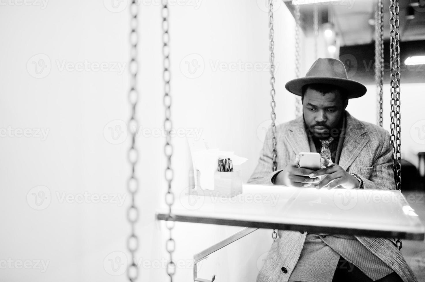 elegante modello uomo afroamericano in cappotto grigio, cravatta giacca e cappello rosso seduto sull'altalena al bar con il telefono cellulare a portata di mano. foto in bianco e nero.