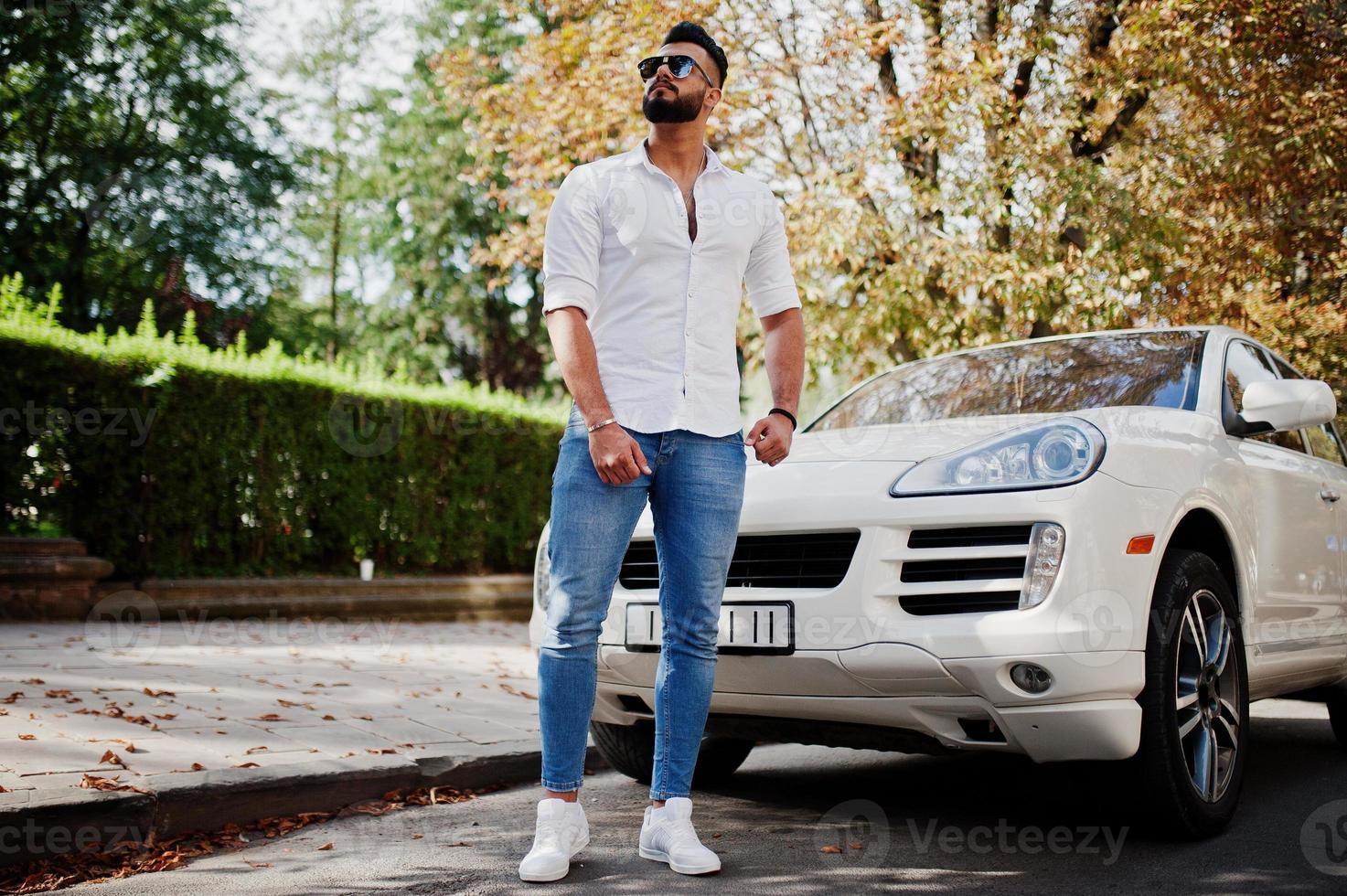 elegante uomo arabo alto modello in camicia bianca, jeans e occhiali da sole posato in una strada della città. ragazzo arabo ricco e attraente della barba contro l'automobile bianca del suv. foto