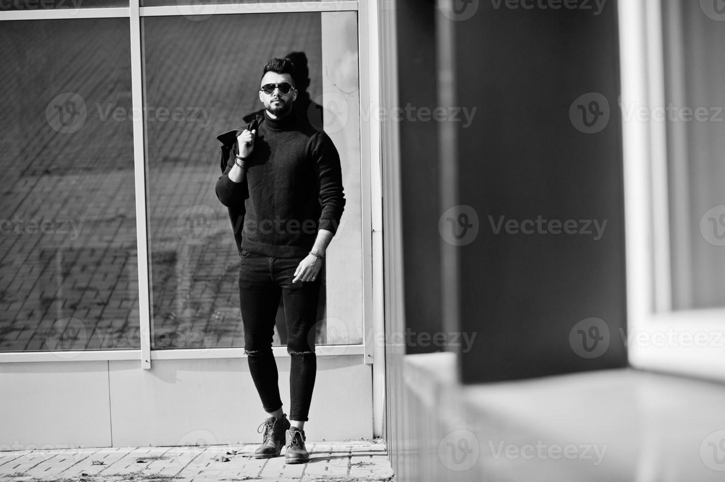 l'uomo arabo con barba ricca di moda indossa il collo alto e gli occhiali da sole posati contro le grandi finestre di un edificio moderno con una giacca a portata di mano. ragazzo modello arabo elegante, di successo e alla moda. foto