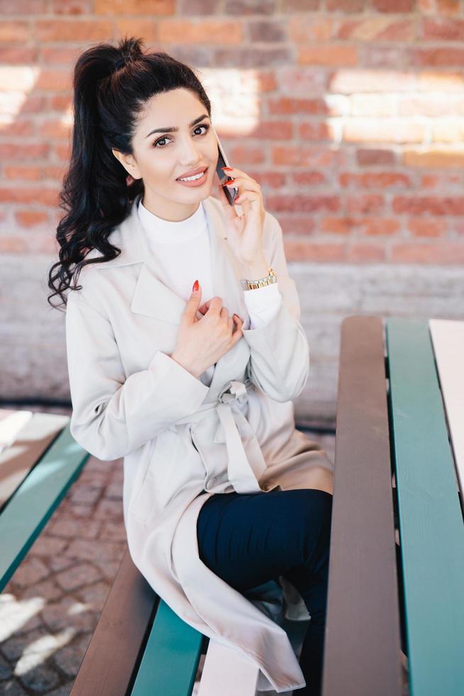 adorabile donna bruna in camice bianco e pantaloni neri seduta al chiuso su un muro di mattoni che parla al cellulare con il suo uomo che discute del loro appuntamento e ricorda momenti piacevoli. concetto di tecnologia foto