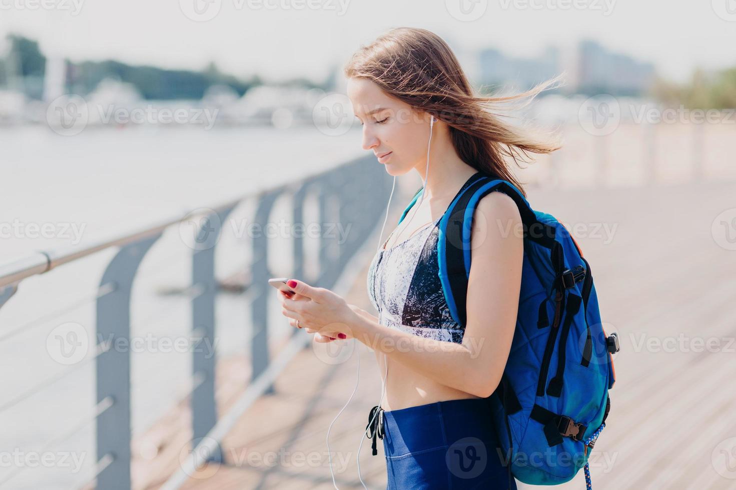 inquadratura laterale di una donna bruna seria guarda seriamente lo schermo dello smartphone, ascolta un libro elettronico, porta lo zaino, posa all'aperto, cammina a lungo, respira aria fresca, gode di un'atmosfera tranquilla foto