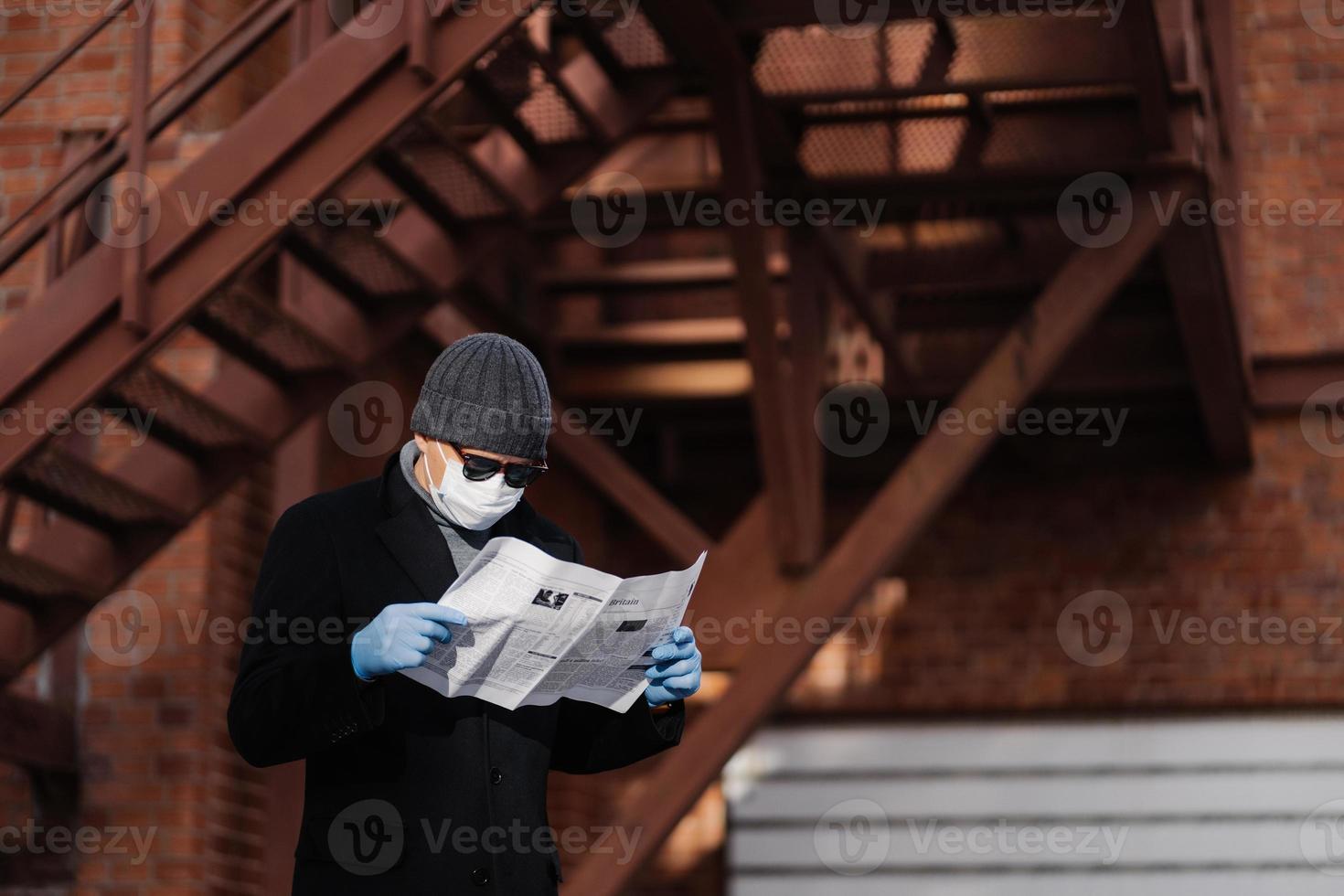 l'uomo concentrato indossa occhiali da sole e cappotto, maschera medica, guanti protettivi in gomma, legge notizie fresche sul coronavirus che si diffondono dai giornali, posa su sfondo sfocato con scale. foto