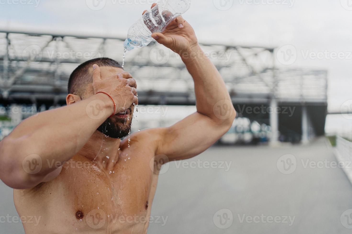 lo sportivo esausto spruzza acqua fresca e fredda sopra la testa, cerca di rinfrescarsi dopo un duro esercizio all'aperto, posa a torso nudo, ha muscoli forti e forma del corpo perfetta. idratazione e concetto di sport foto