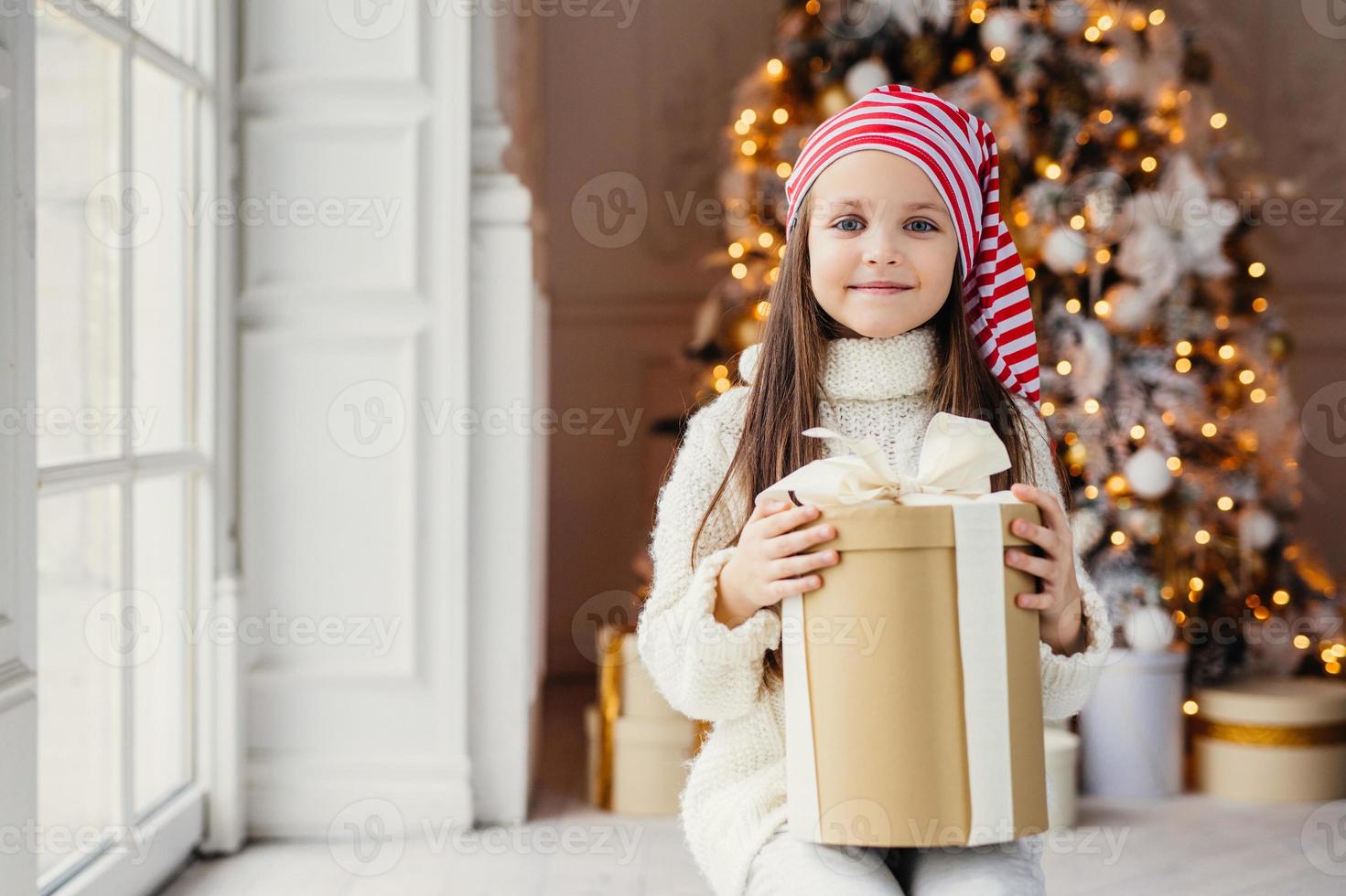 tiro al coperto di un bambino dall'aspetto piacevole con occhi azzurri affascinanti, indossa un cappello da Babbo Natale, tiene il regalo in una scatola avvolta, si siede su uno sfondo decorato di Capodanno. infanzia, concetto di celebrazione foto