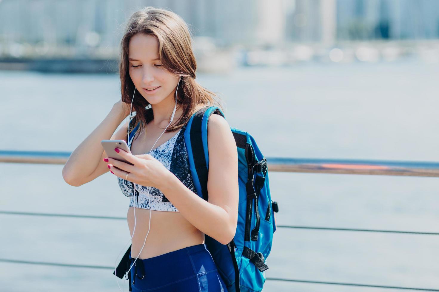 ritratto orizzontale di una bella donna con una figura perfetta, indossa leggings e top, ha uno sguardo concentrato sullo smartphone, porta un grande zaino, cerca di trovare il percorso corretto mentre copre lunghe distanze da solo foto