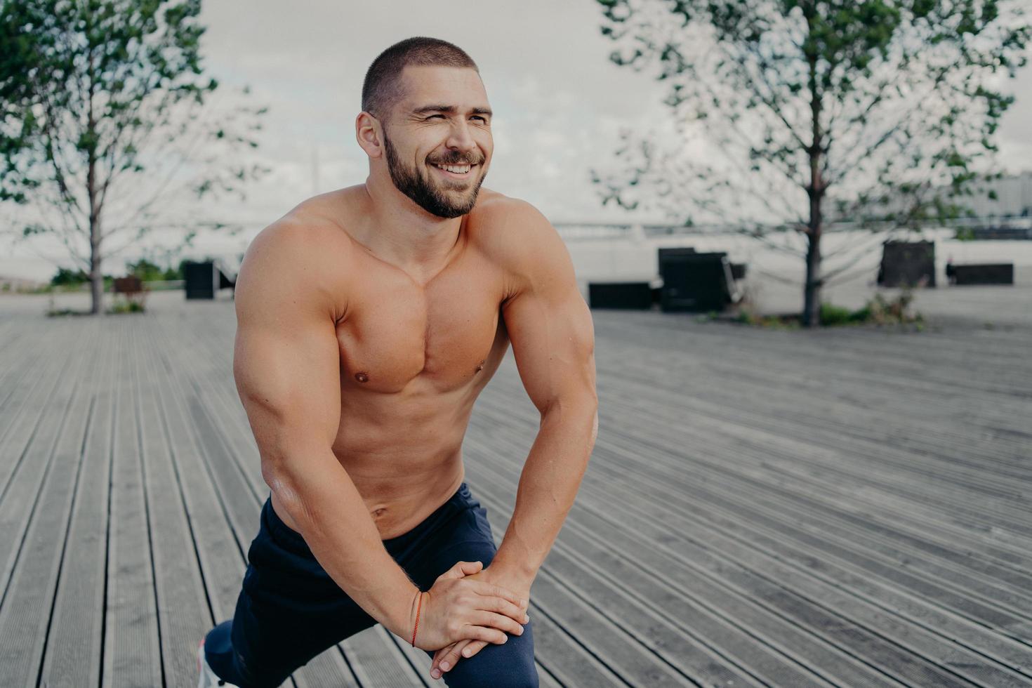 concetto di routine mattutina quotidiana ed esercizi sportivi. il bodybuilder sportivo con la barba lunga con l'espressione allegra fa esercizi di stretching per le gambe, posa con il busto nudo, si allena all'aperto. foto