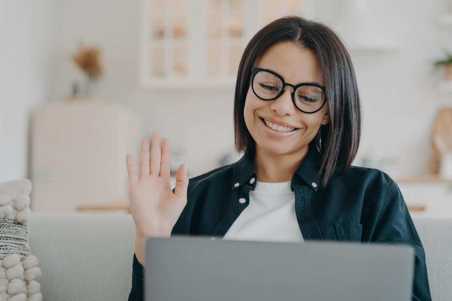 la ragazza imprenditrice con gli occhiali ha una videochiamata sul computer e agitando la mano. lavoro a distanza a casa. foto