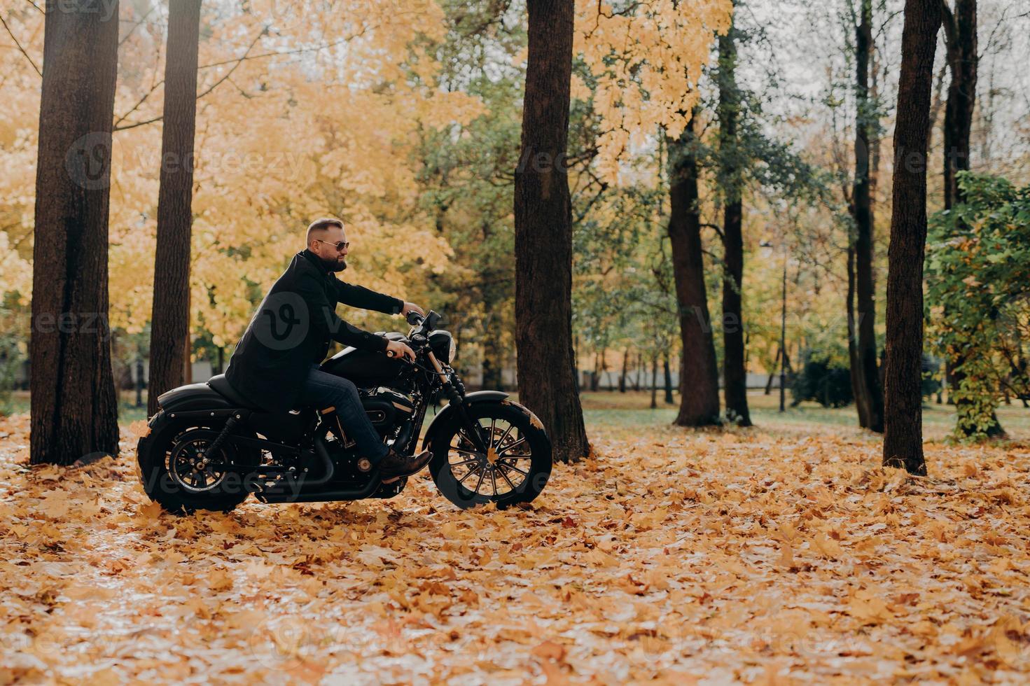 bel motociclista barbuto spensierato va in moto veloce, indossa occhiali da sole, va in bicicletta nel parco, posa contro alberi e fogliame gialli, spazio vuoto per le tue informazioni. turistico all'aperto foto