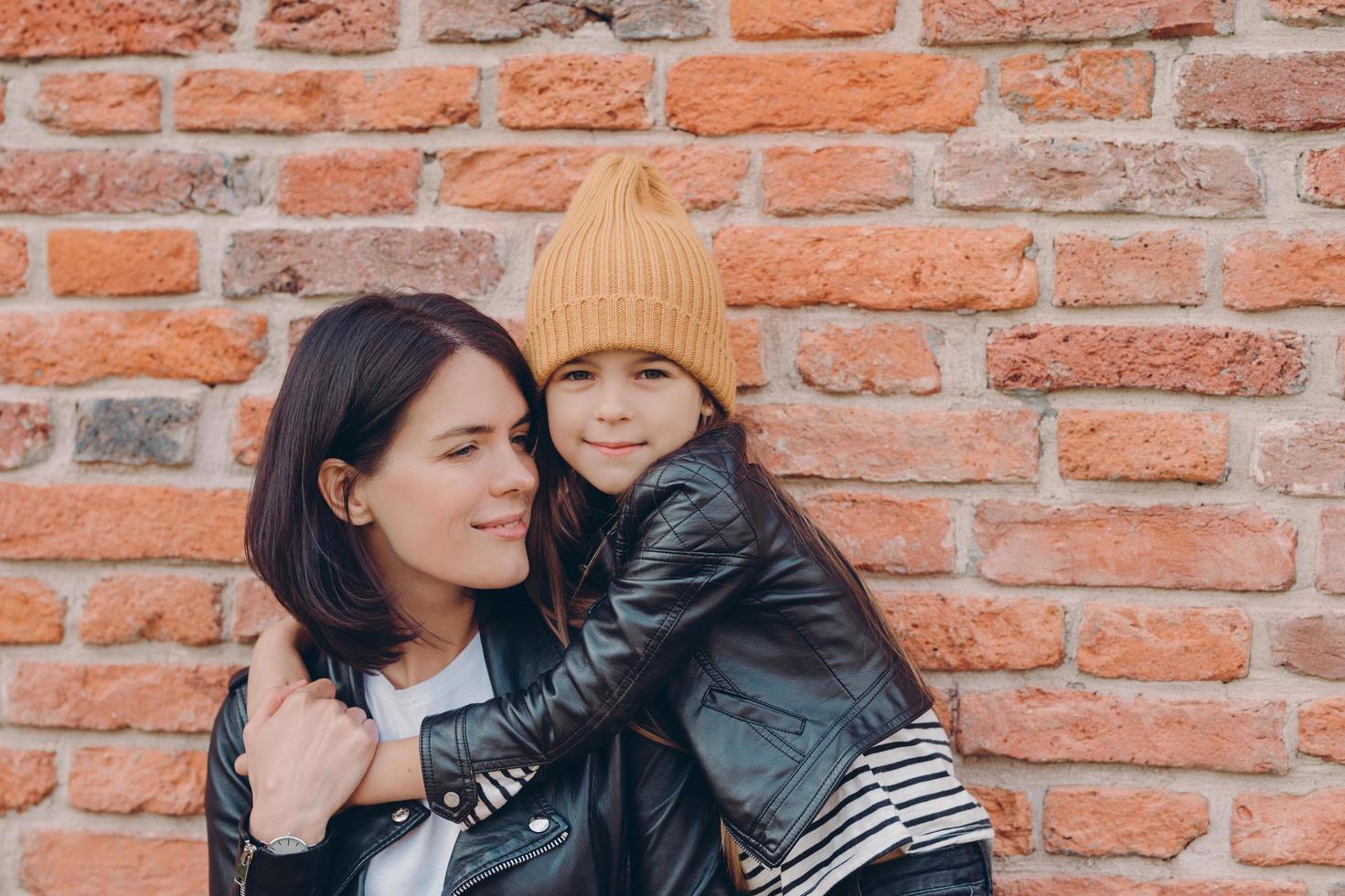 la madre affettuosa con i capelli scuri riceve un caldo abbraccio dalla figlia, indossa giacche di pelle, posa insieme contro un muro di mattoni. la bambina alla moda con il cappello abbraccia sua madre. concetto di famiglia e bambini foto