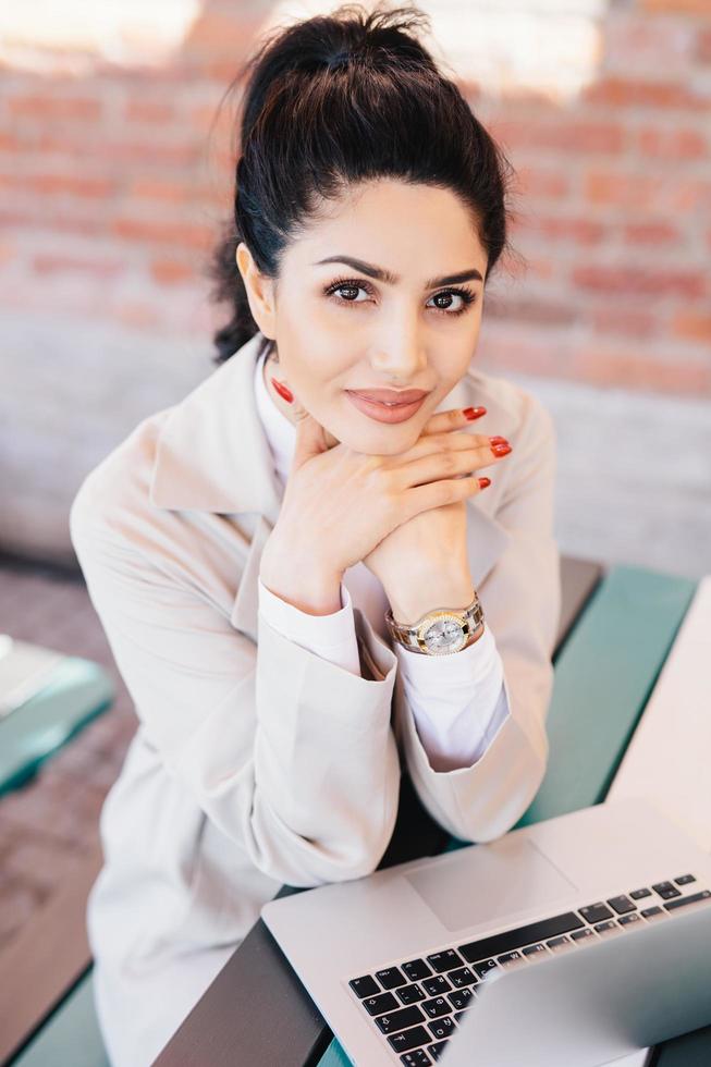 giovane donna d'affari bruna con occhi affascinanti, mani gentili con manicure rossa che indossa un orologio a portata di mano e camice bianco che si tiene per mano sotto il mento seduta vicino al suo computer portatile che si riposa dopo un duro lavoro foto