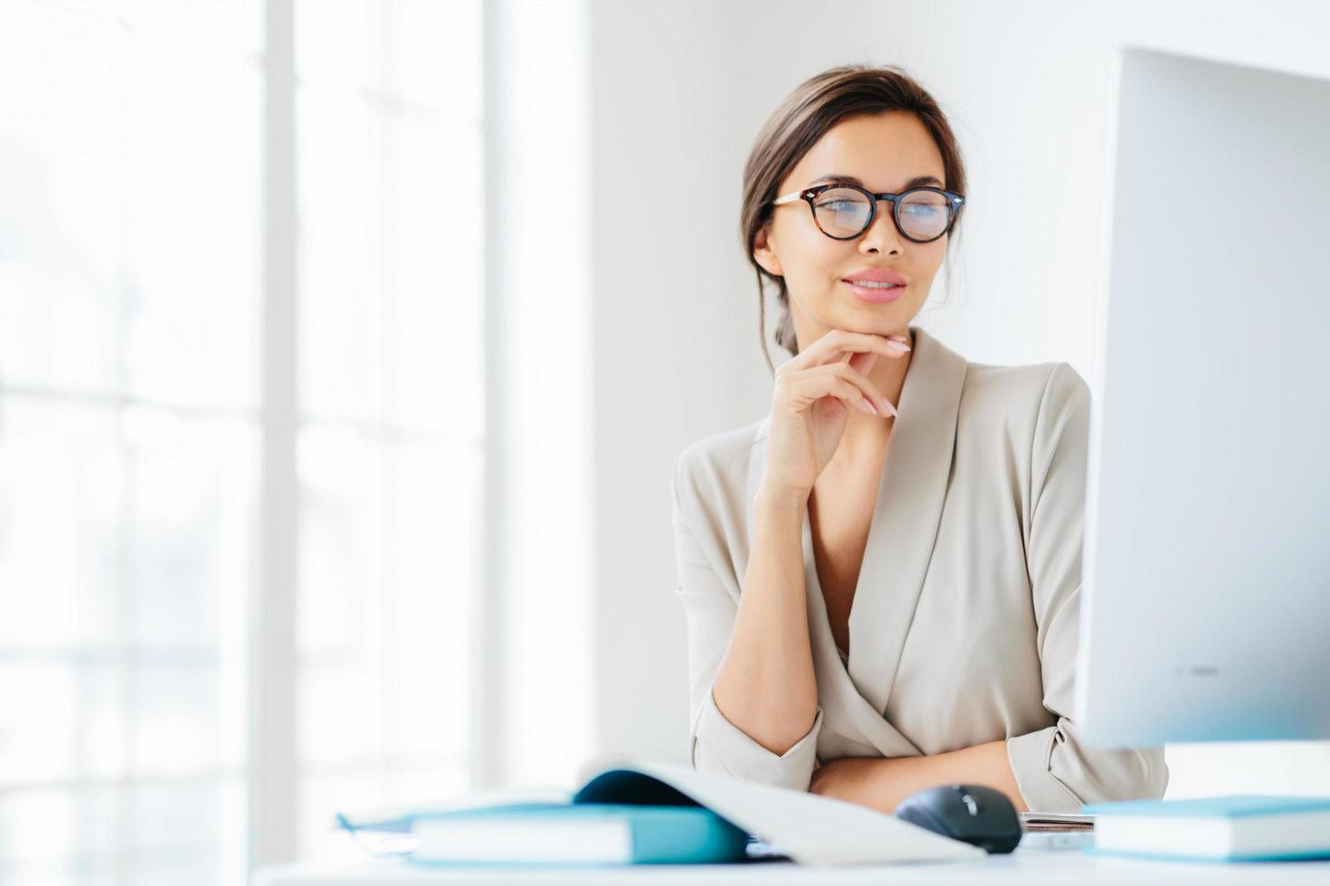 affascinante impiegato femminile tiene la mano sotto il mento, guarda sullo schermo del computer, indossa occhiali per la correzione della vista, posa nel proprio gabinetto, prepara le informazioni per futuri incontri di lavoro foto