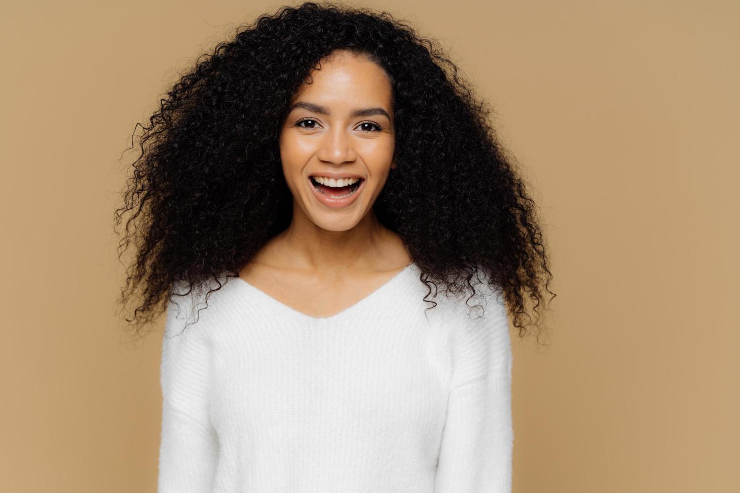 colpo alla testa di un modello femminile sano e positivo ride felice, ha folti capelli afro, vestito con un maglione morbido bianco, felice di ricevere complimenti dal fidanzato, isolato su sfondo marrone. concetto di emozioni foto