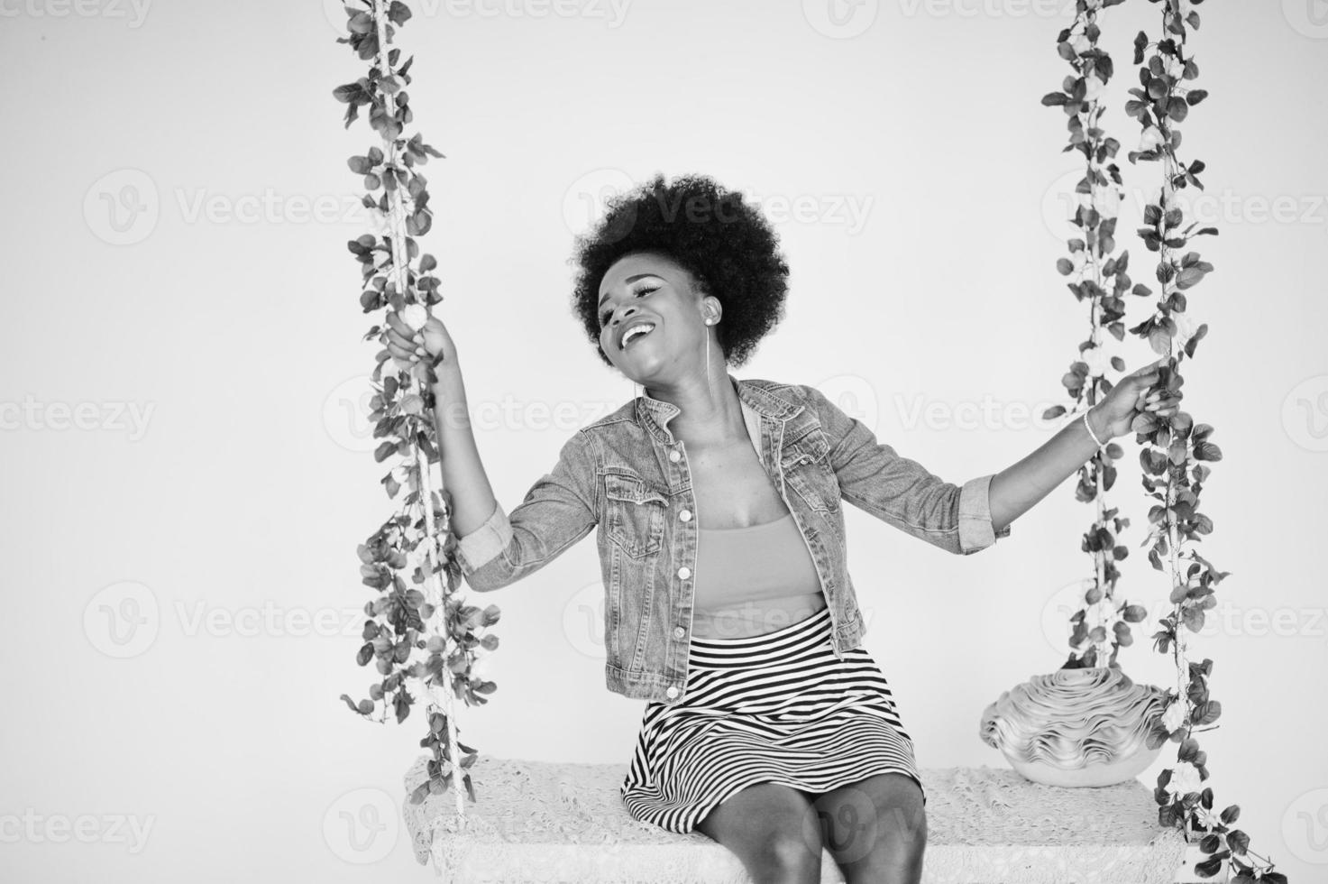 attraente donna afroamericana con capelli afro su gonna e giacca di jeans, posata in una stanza bianca sull'altalena. modello nero alla moda. foto