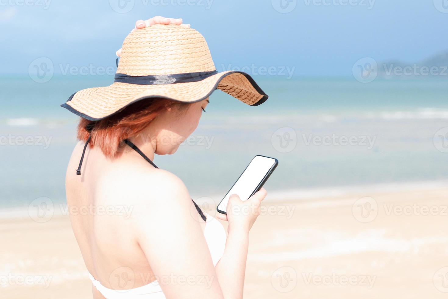 una donna che indossa un costume da bagno e un cappello da sole sta utilizzando un telefono cellulare in piedi vicino alla spiaggia. cellulare vuoto per mock up. foto