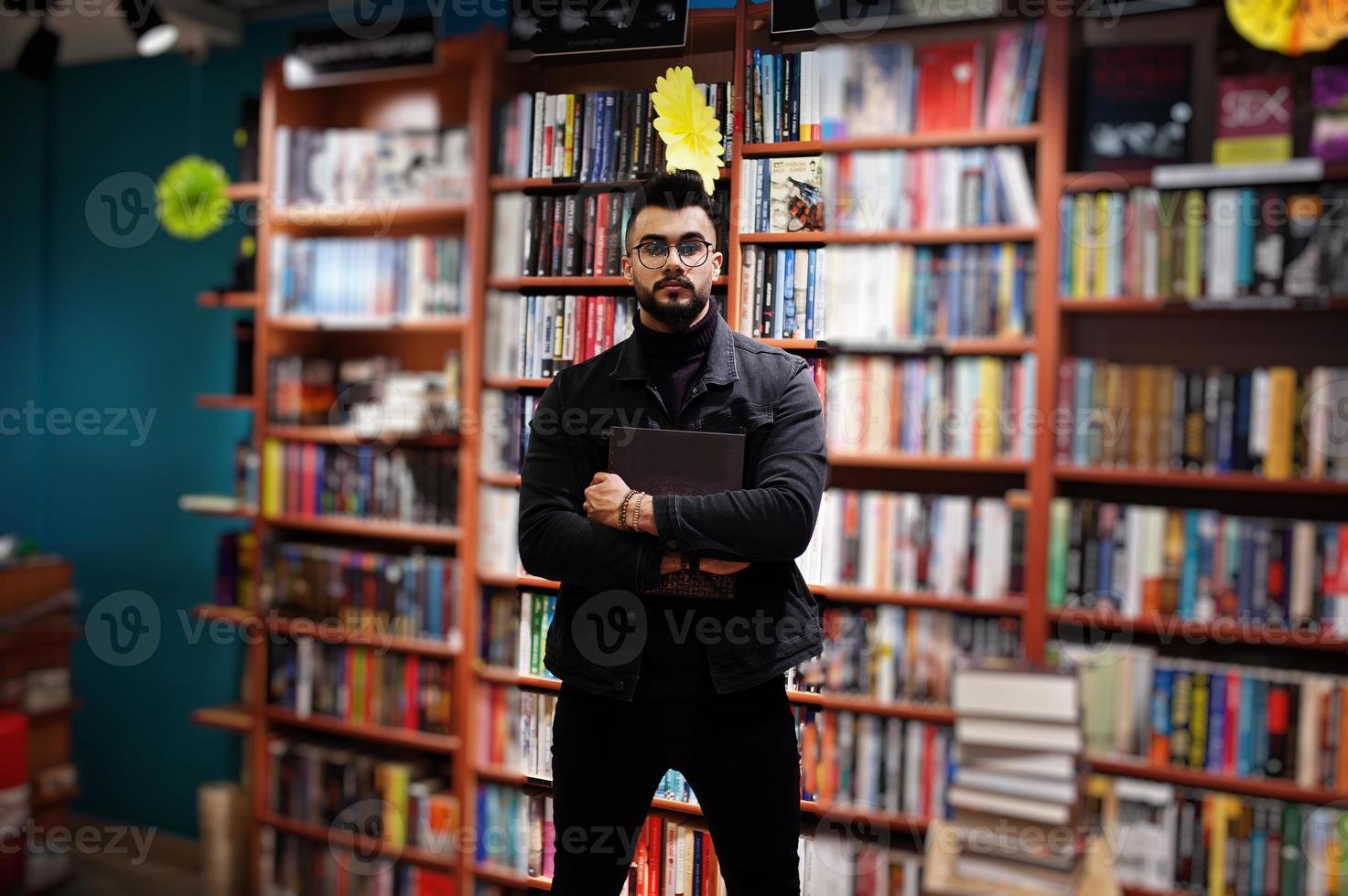 studente arabo alto e intelligente, indossa una giacca di jeans nera e occhiali da vista, in biblioteca con un libro a portata di mano. foto