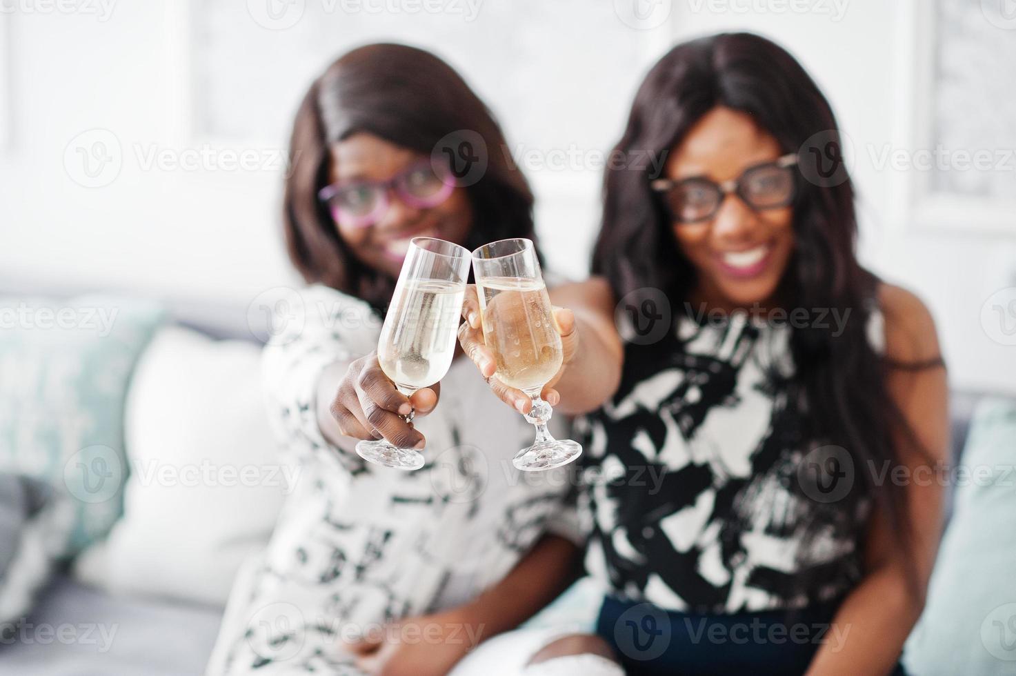 due amiche africane indossano occhiali in posa in una stanza bianca interna e bevono champagne dai bicchieri. foto