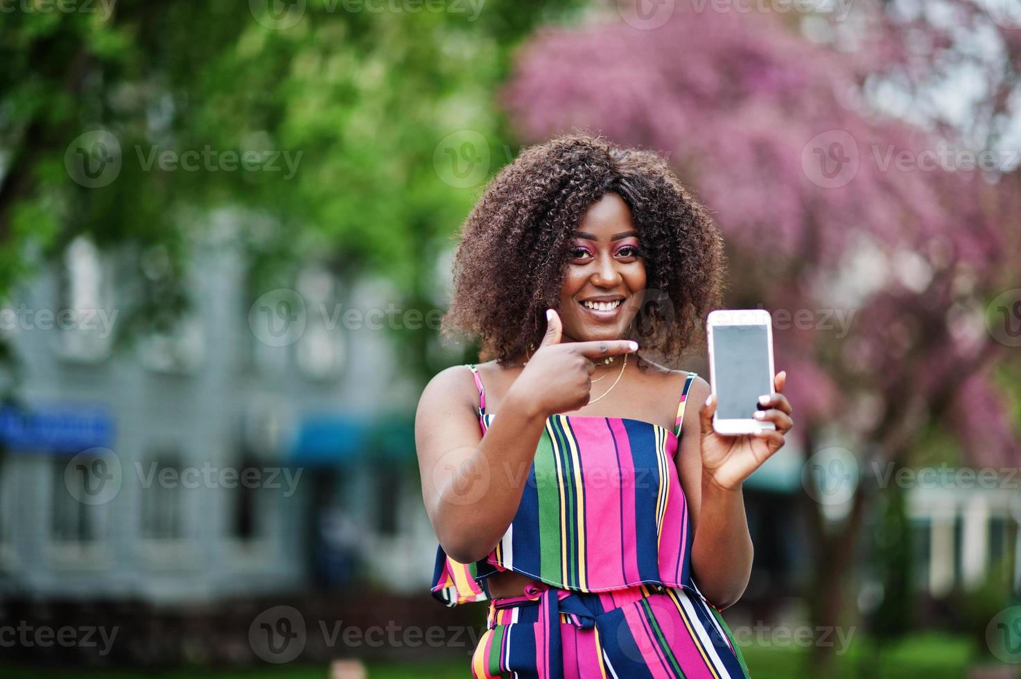 donna afroamericana alla moda in tuta a righe rosa posata in strada in fiore primaverile e mostra il dito sullo schermo del telefono cellulare. foto