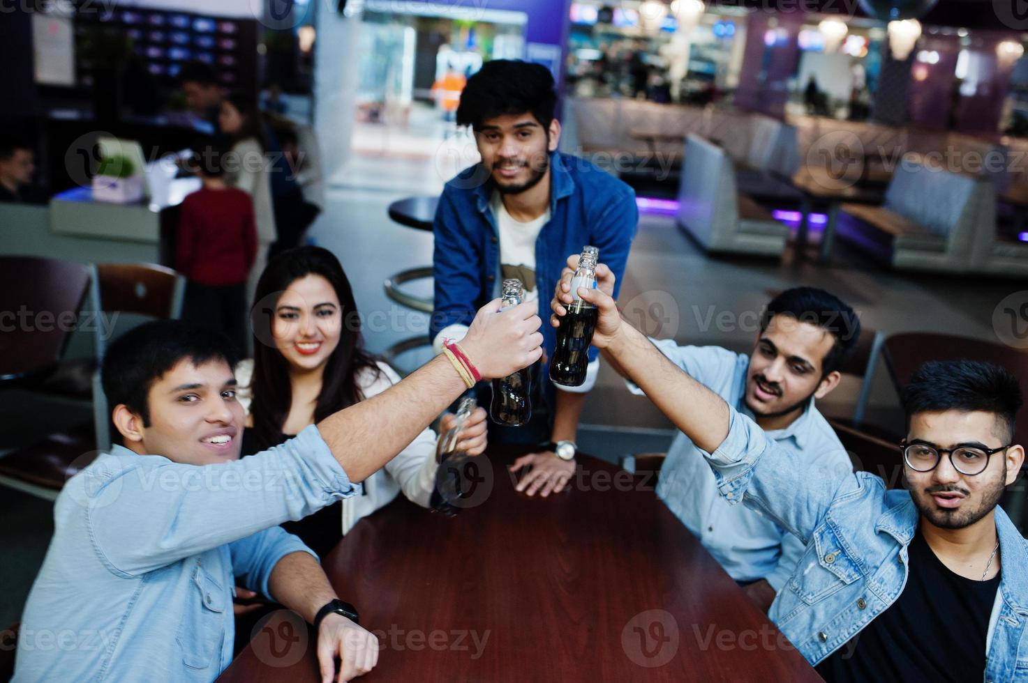 gruppo di amici asiatici alla moda indossa jeans seduti a tavola e tifa con bottiglie di soda nel club. foto