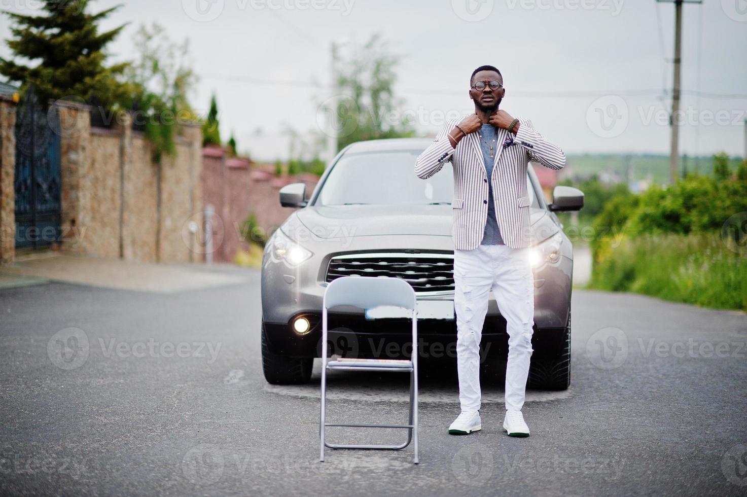 uomo afroamericano ricco ed elegante in giacca e pantaloni bianchi, occhiali posati contro un'auto suv. foto