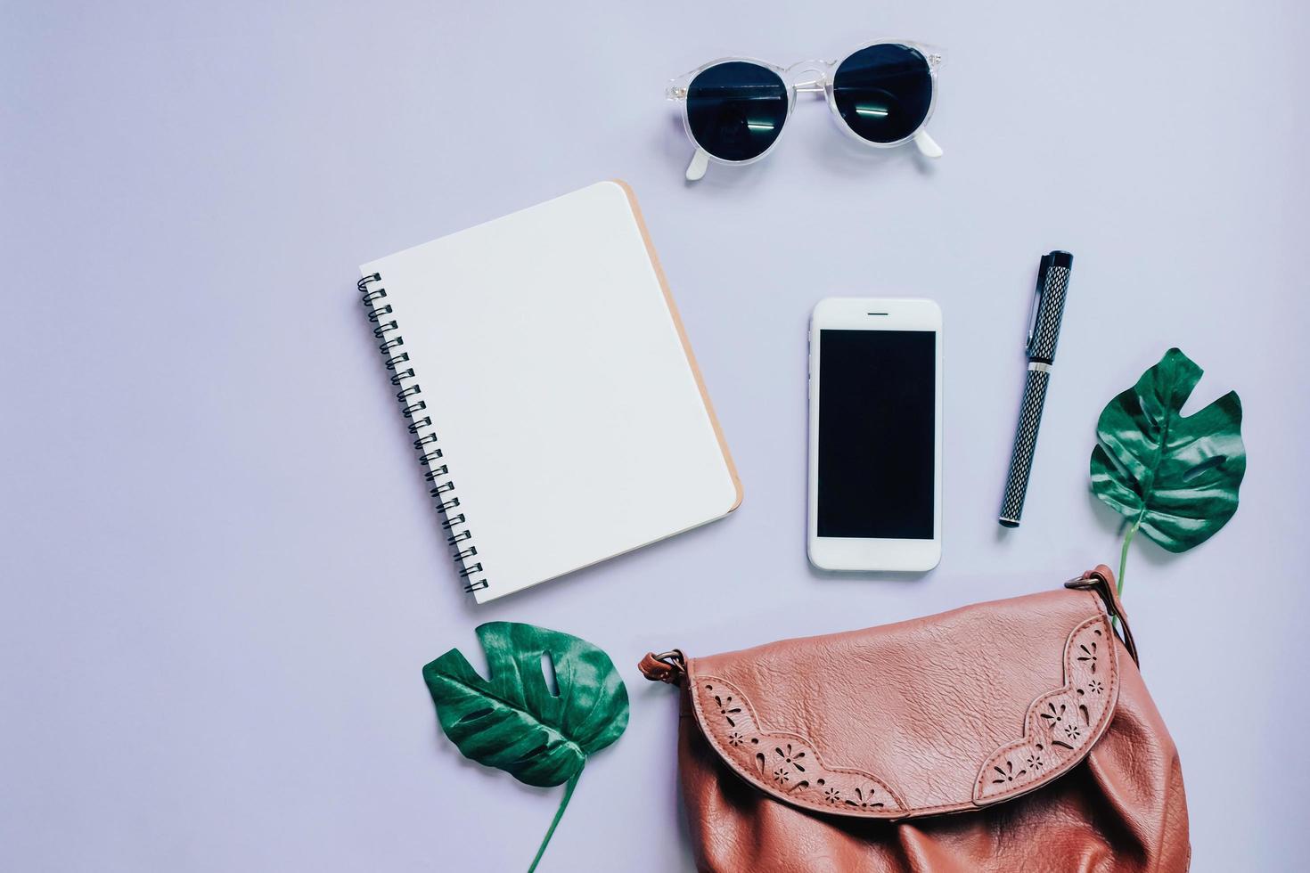 vista dall'alto piatto di borsa da donna con smartphone, taccuino bianco, occhiali da sole e foglia verde, spazio per la copia foto