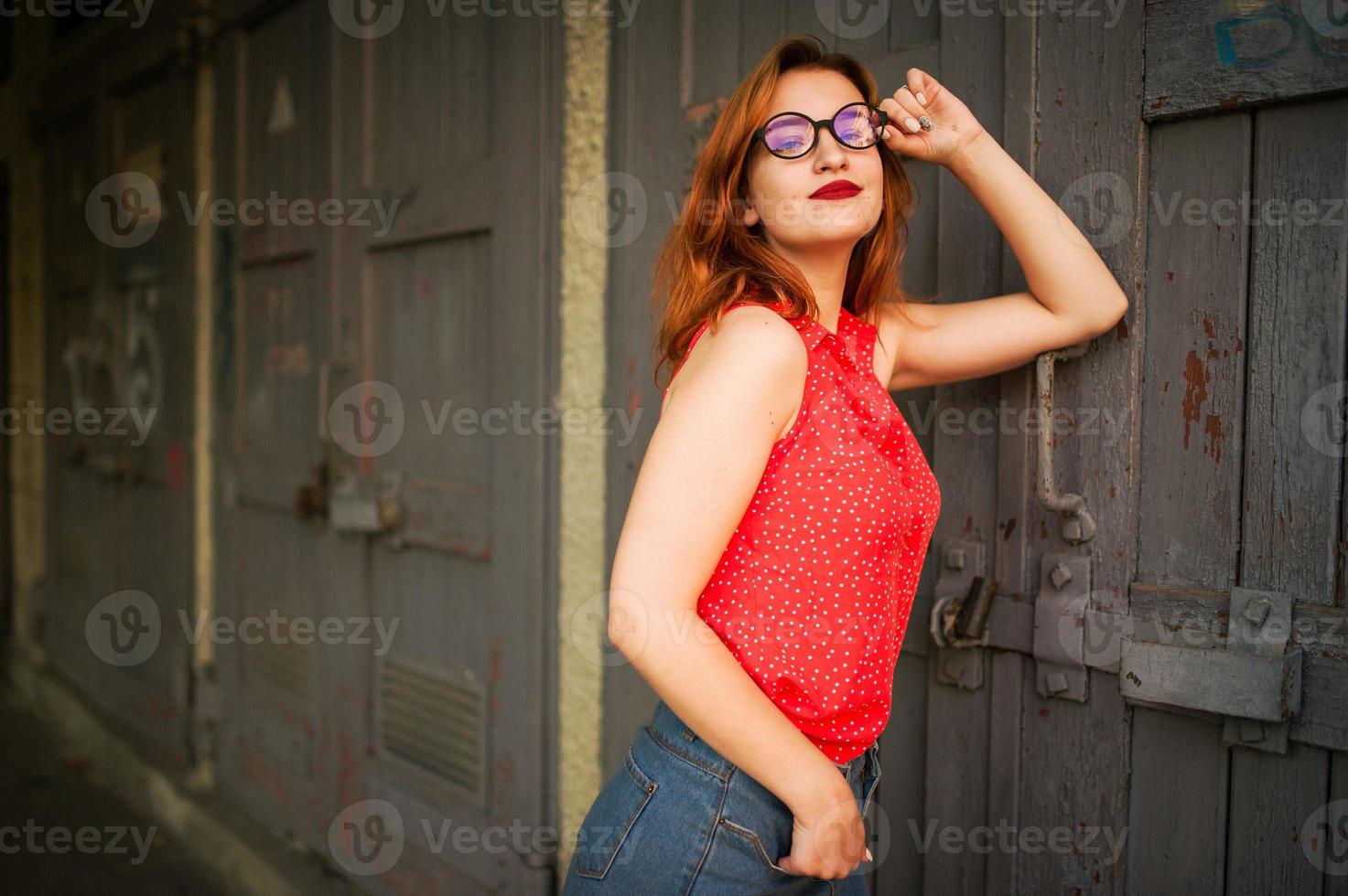 attraente donna dai capelli rossi in occhiali, indossare su camicetta rossa e gonna di jeans in posa. foto