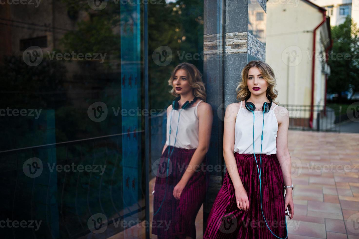 ragazza modello bionda alla moda e bella in elegante gonna di velluto di velluto rosso, camicetta bianca, posata con telefono e auricolari contro le finestre dell'edificio della città. foto