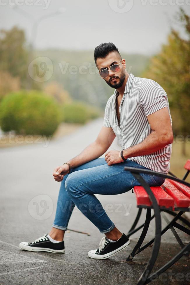 l'uomo alla moda con barba araba alta indossa camicia, jeans e occhiali da sole seduto su una panchina rossa al parco. foto