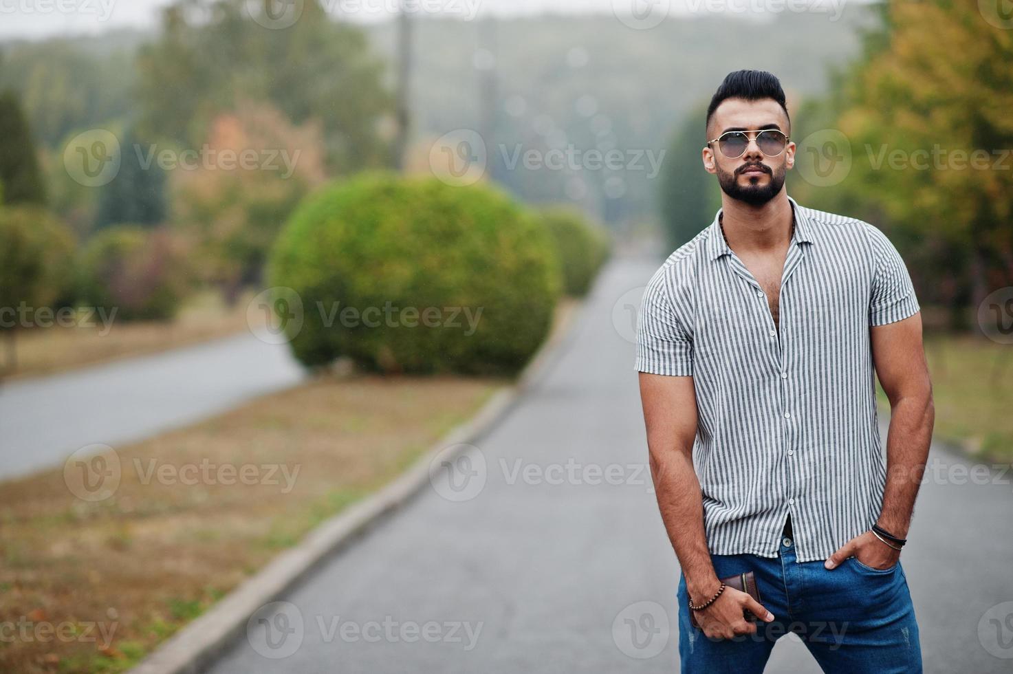 l'uomo alla moda con barba araba alta indossa camicia, jeans e occhiali da sole in posa sul parco e tiene il portafoglio a portata di mano. foto