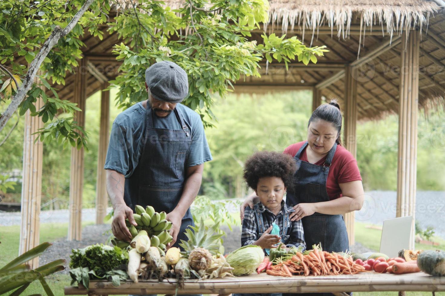 la famiglia felice di agricoltori della diversità, il papà nero e la mamma asiatica con il figlio in grembiule vendono insieme prodotti naturali dell'orto, verdure biologiche, frutta e attività del mercato agricolo locale in campagna. foto