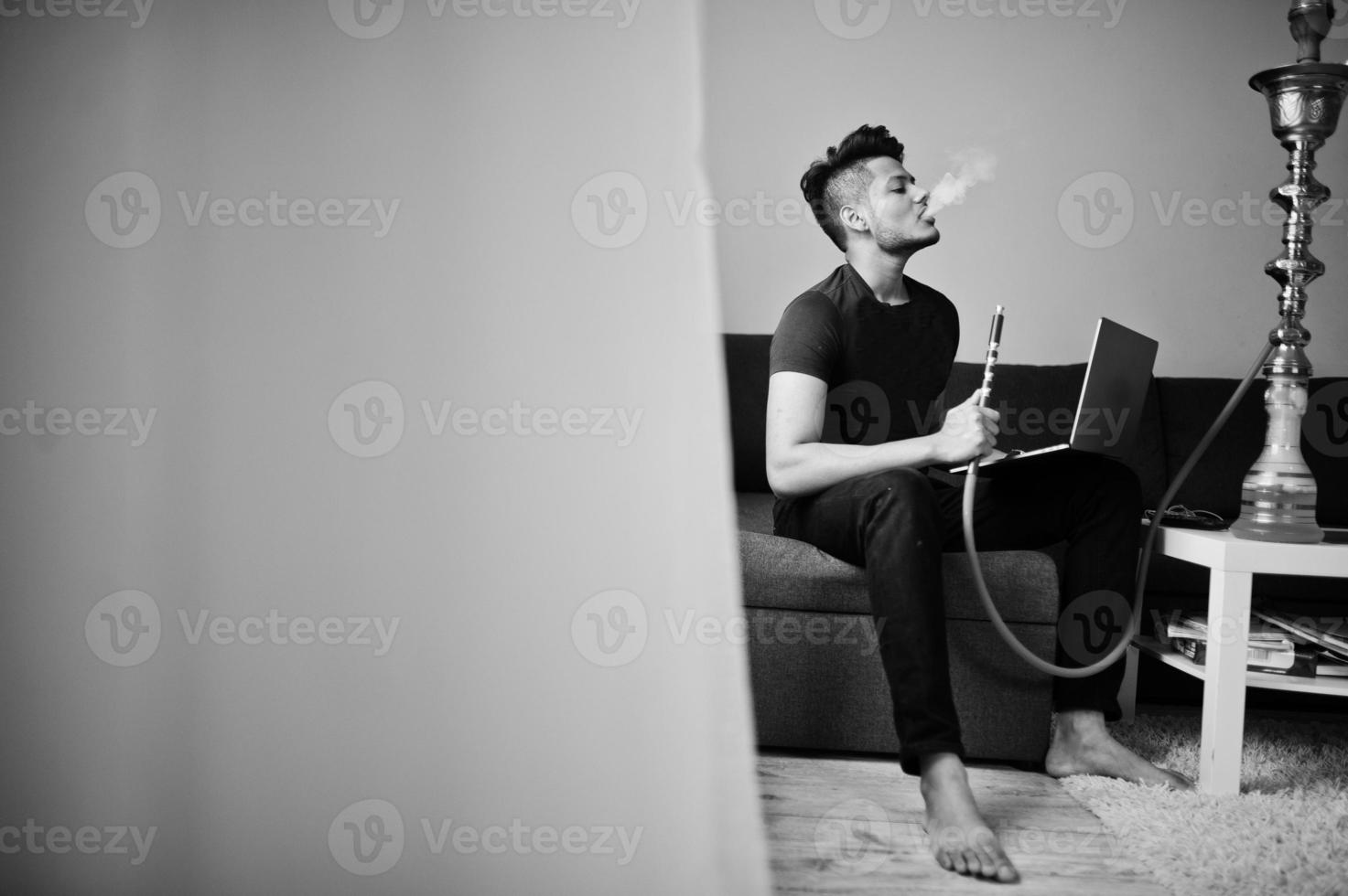 uomo indiano bello e alla moda in nero seduto in camera, fumare narghilè e lavorare al computer portatile. foto