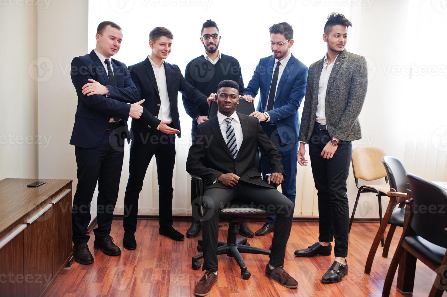 sei uomini d'affari multirazziali in piedi in ufficio e rotolano l'uomo sulla sedia. gruppo eterogeneo di dipendenti maschi in abiti formali che si divertono. foto