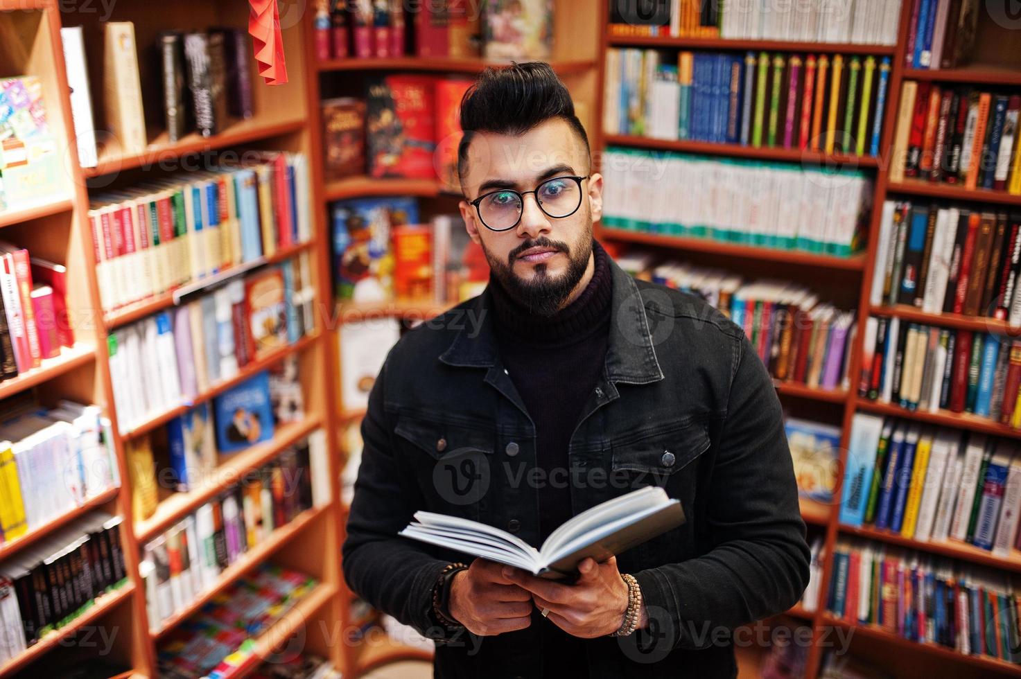 studente arabo alto e intelligente, indossa una giacca di jeans nera e occhiali da vista, in biblioteca con un libro a portata di mano. foto