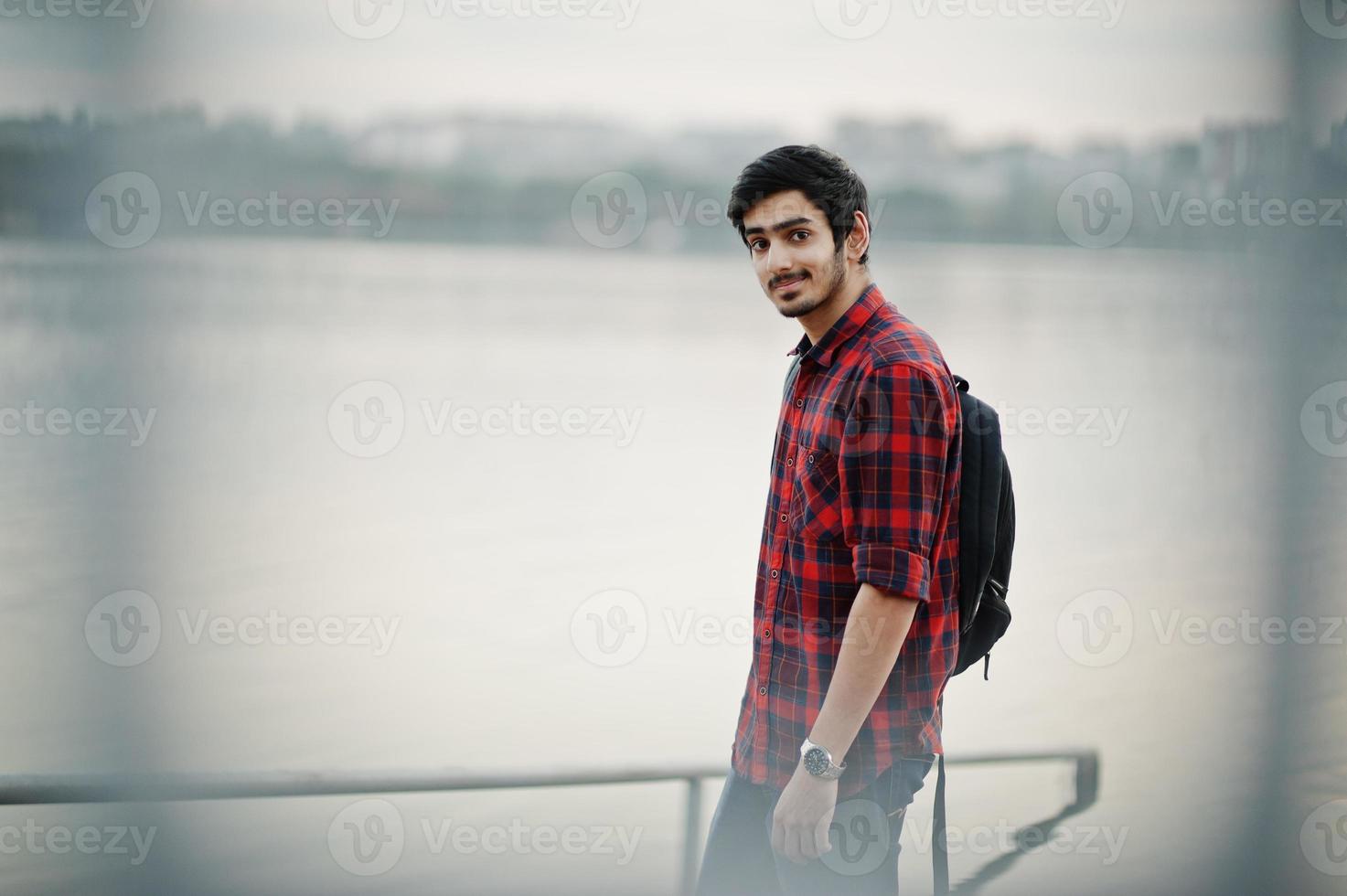 giovane studente indiano in camicia a scacchi e jeans con zaino in posa sulla città di sera contro il lago. foto