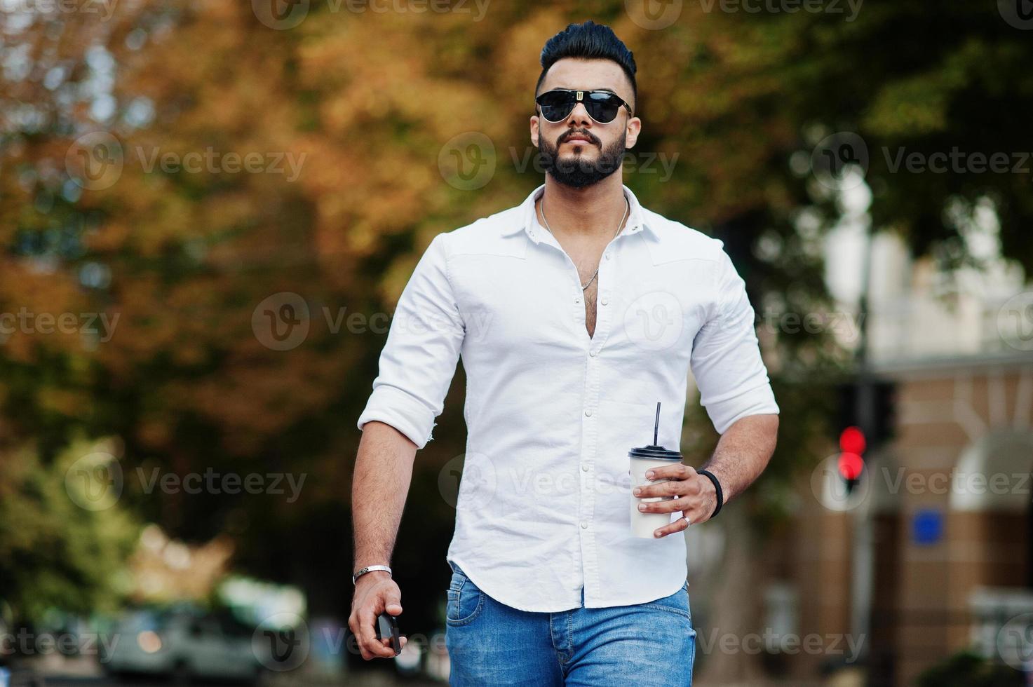 elegante uomo arabo alto modello in camicia bianca, jeans e occhiali da sole posato in una strada della città. barba attraente ragazzo arabo con una tazza di caffè a piedi. foto