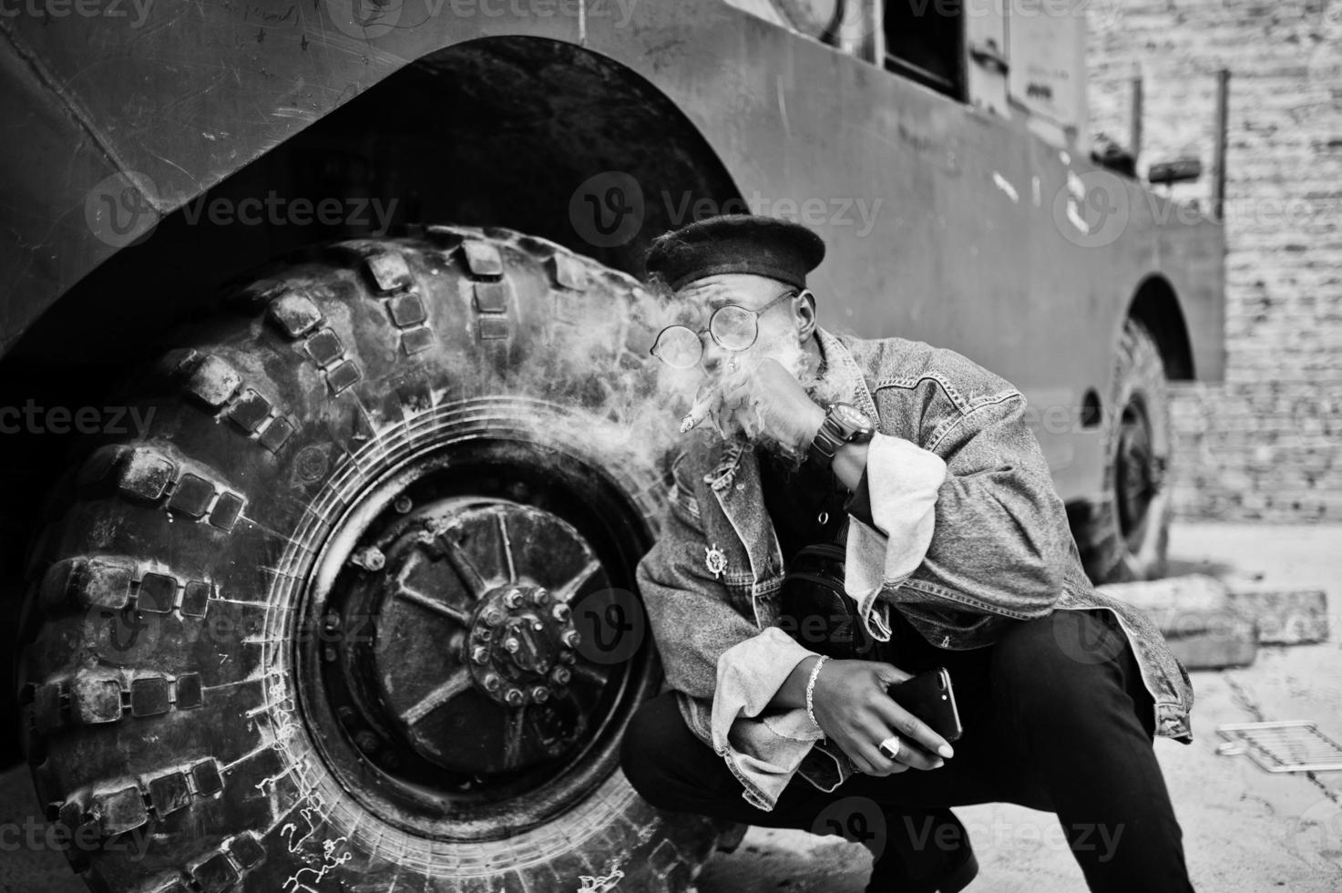 uomo afroamericano in giacca di jeans, berretto e occhiali da vista, fumando sigaro e posato contro la ruota del veicolo blindato militare btr. foto